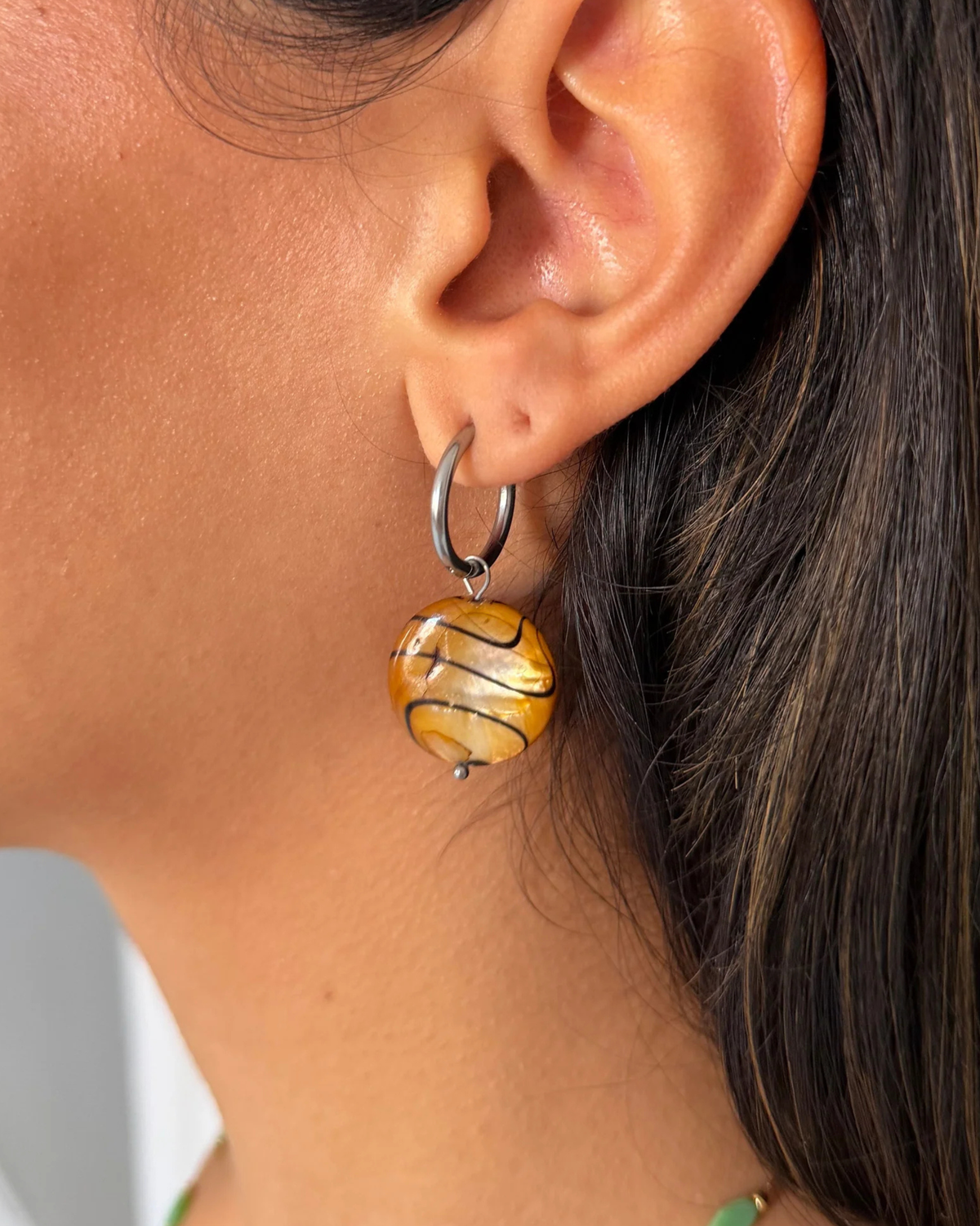 Close-up of a woman's ear wearing a silver huggie hoop earring with a dangling marbled yellow, gold, and brown glass drop earring.