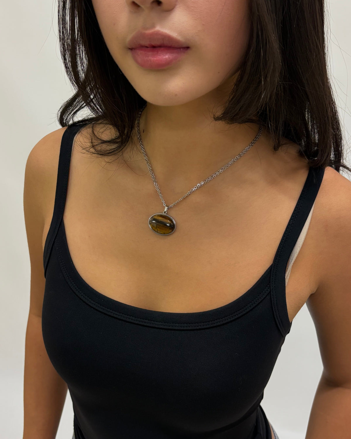 A close-up of a woman wearing a gold chain necklace with an oval tiger's eye pendant, paired with a black tank top.