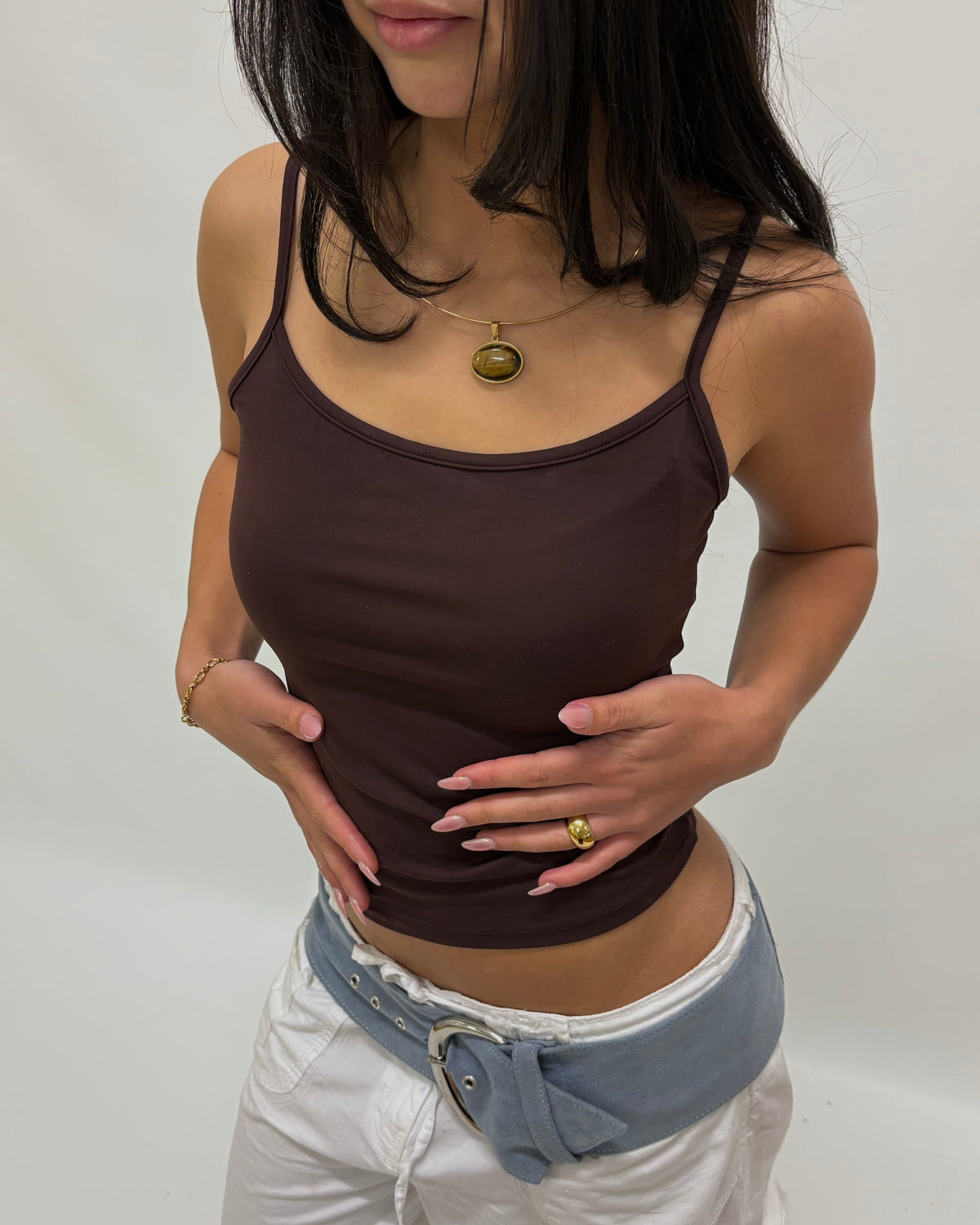A close-up of a model wearing a delicate gold wire necklace with an oval pendant containing a yellow-green and black pattern, tigers eye stone paired with gold hoop earrings and a brown top.