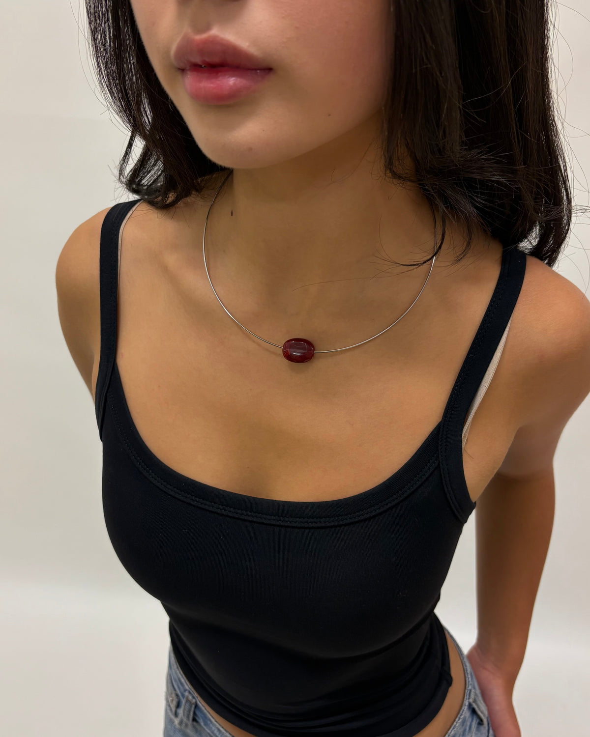 A close-up of a woman wearing a thin silver necklace with a small oval dark red pendant, paired with a black camisole top and light-wash denim jeans.