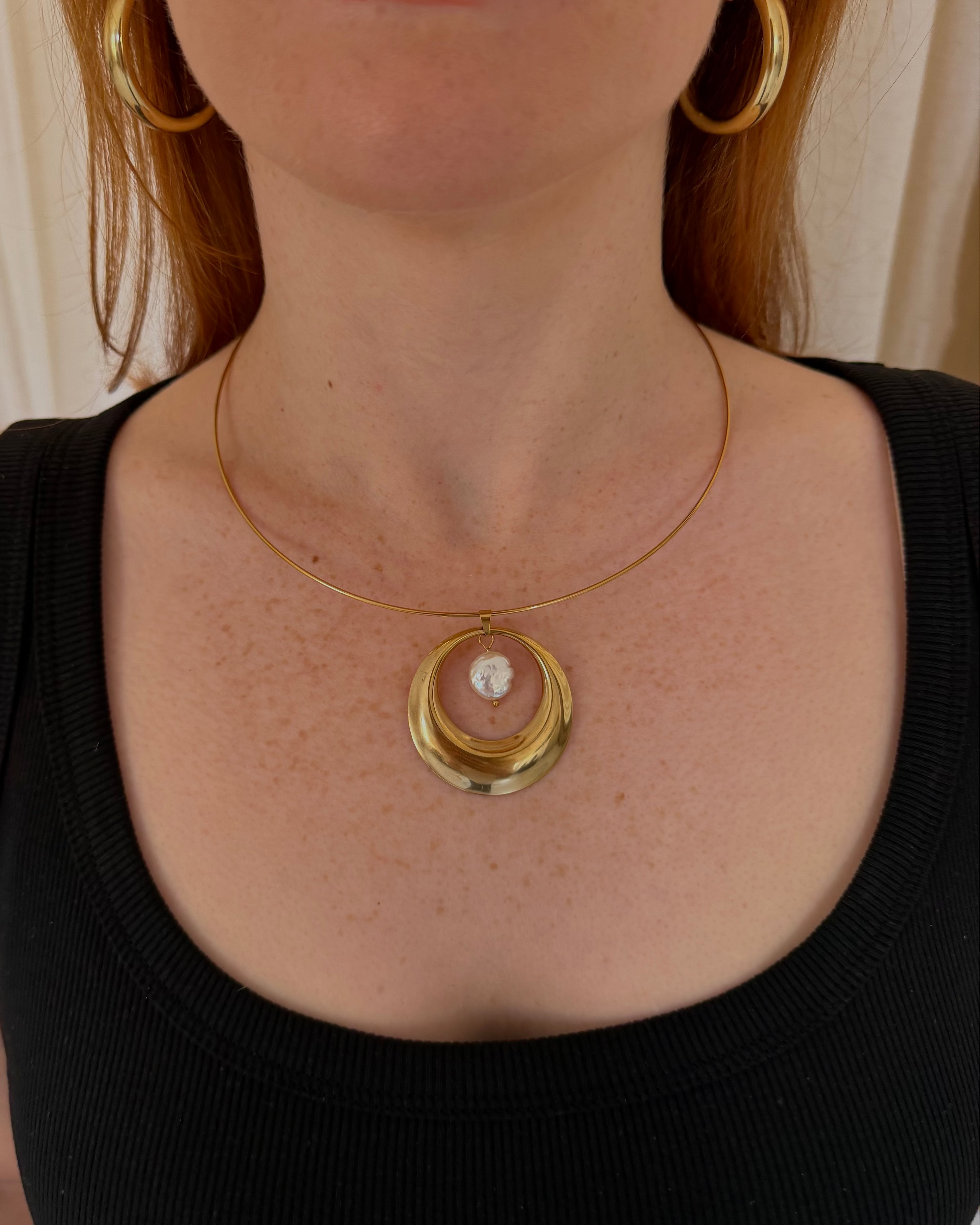 A close-up of a woman wearing a gold necklace with a circular pendant and matching gold hoop earrings, paired with a black ribbed tank top.