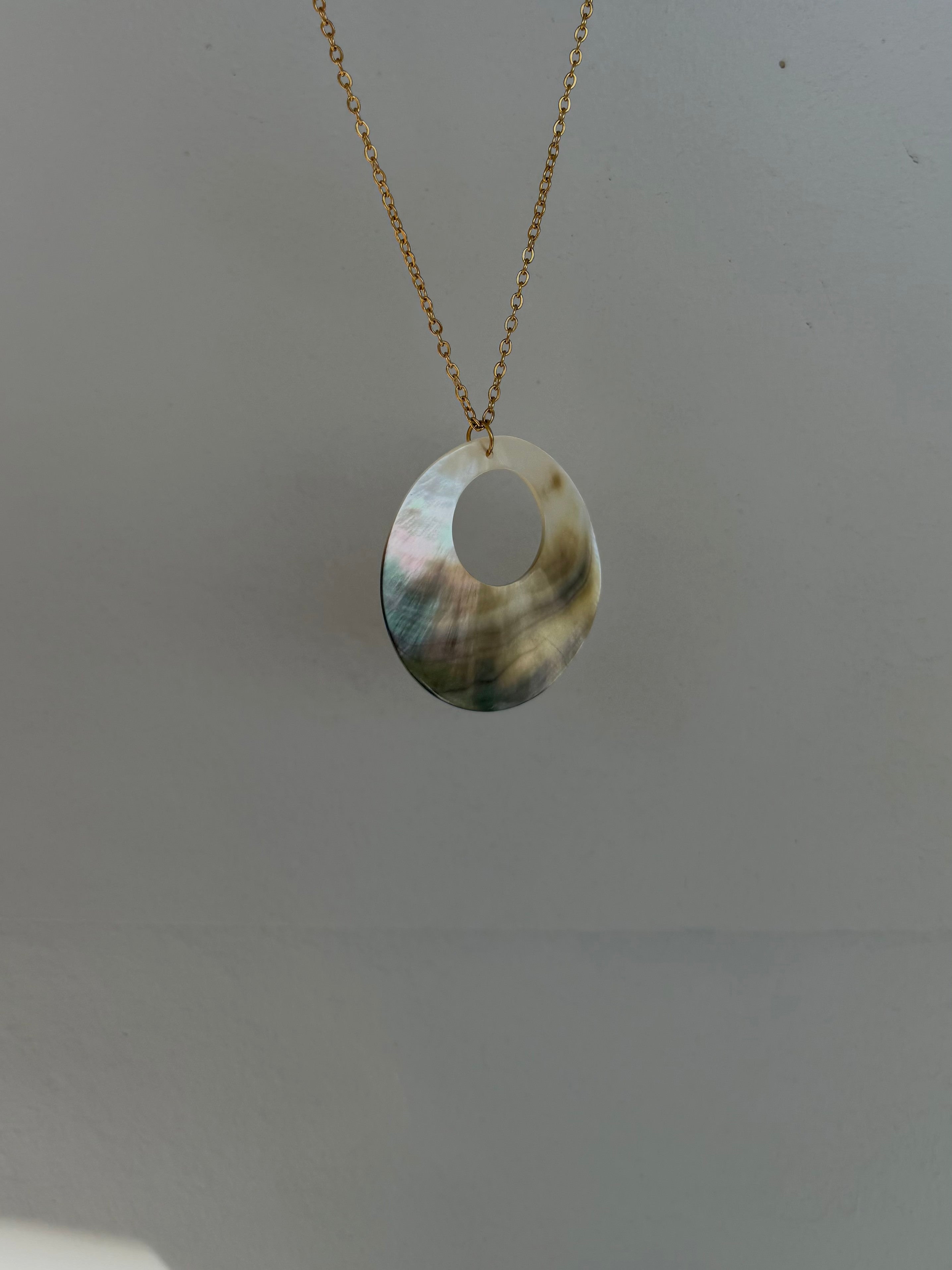 A gold chain necklace with a large, irregularly shaped oval pendant made of iridescent abalone shell, hanging against a plain light gray background.
