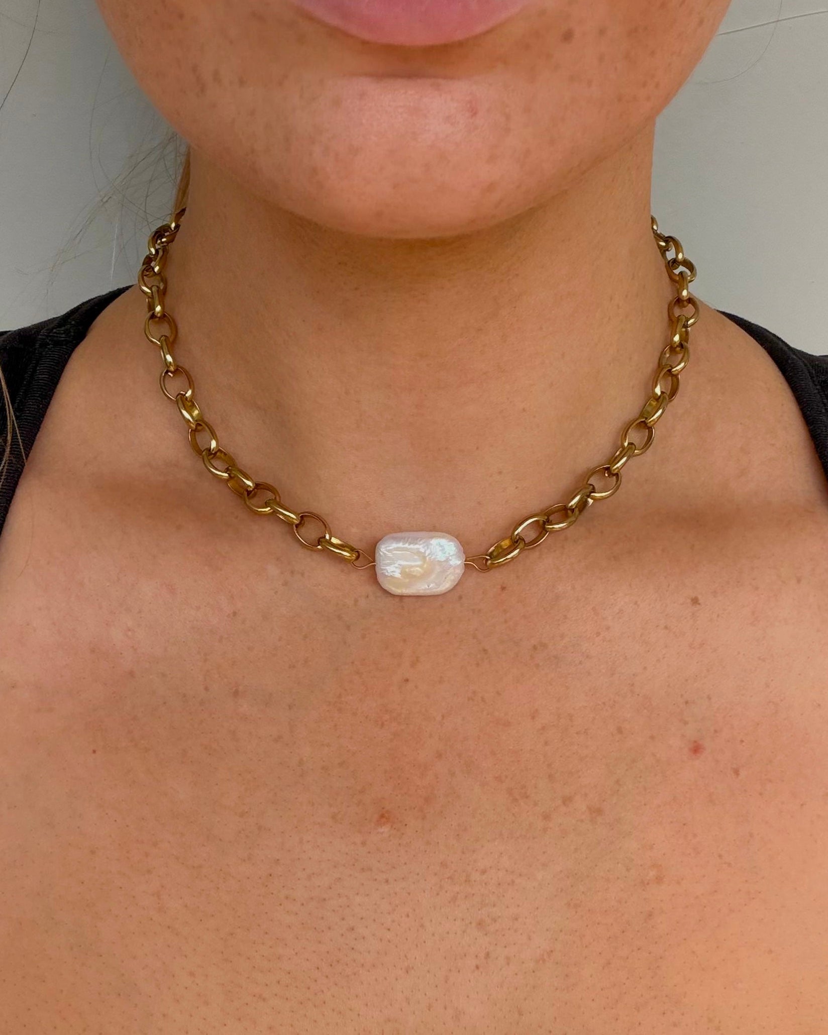 A close-up of a model wearing a gold chain necklace with a central white baroque shell pendant.
