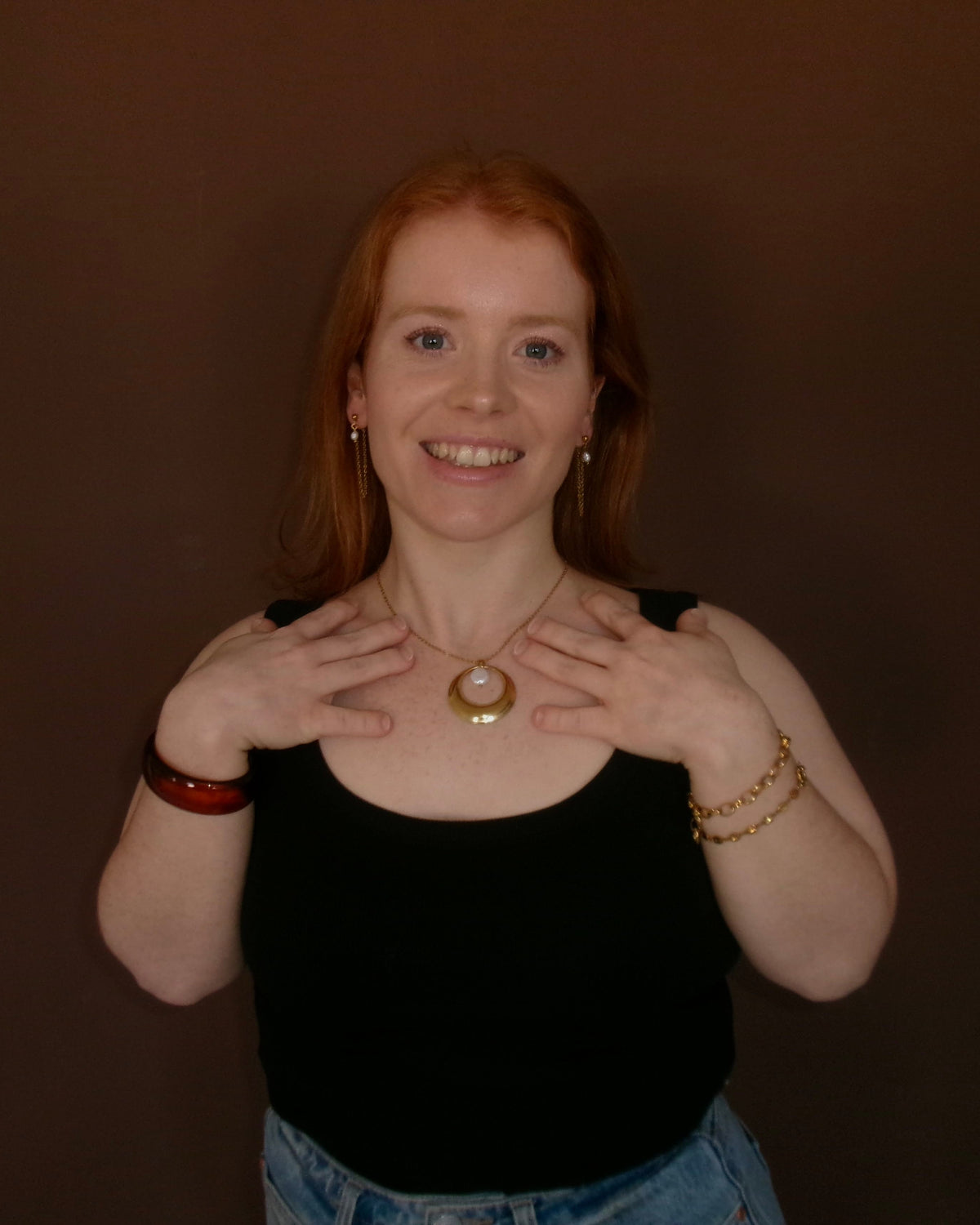 A smiling young woman with red hair wearing a black tank top and blue jeans, holding her hands to her chest to display a gold necklace with a round white pendant. She is standing against a solid dark brown background.