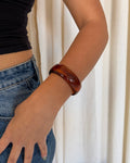 A close-up of a person wearing a glossy, dark reddish-brown wooden bangle bracelet on their wrist, paired with a black crop top and blue jeans, set against a soft cream curtain background.