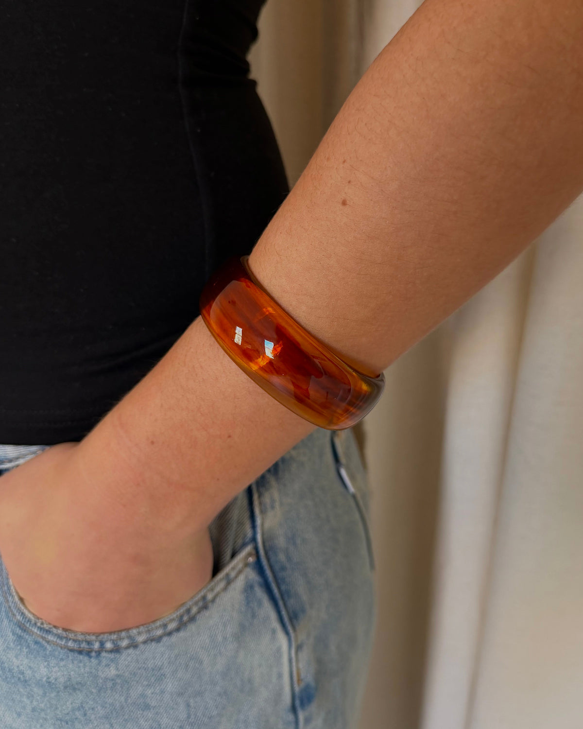 A close-up of a person's forearm wearing a thick, translucent amber-brown resin bangle bracelet, paired with a black top and light-wash blue jeans.