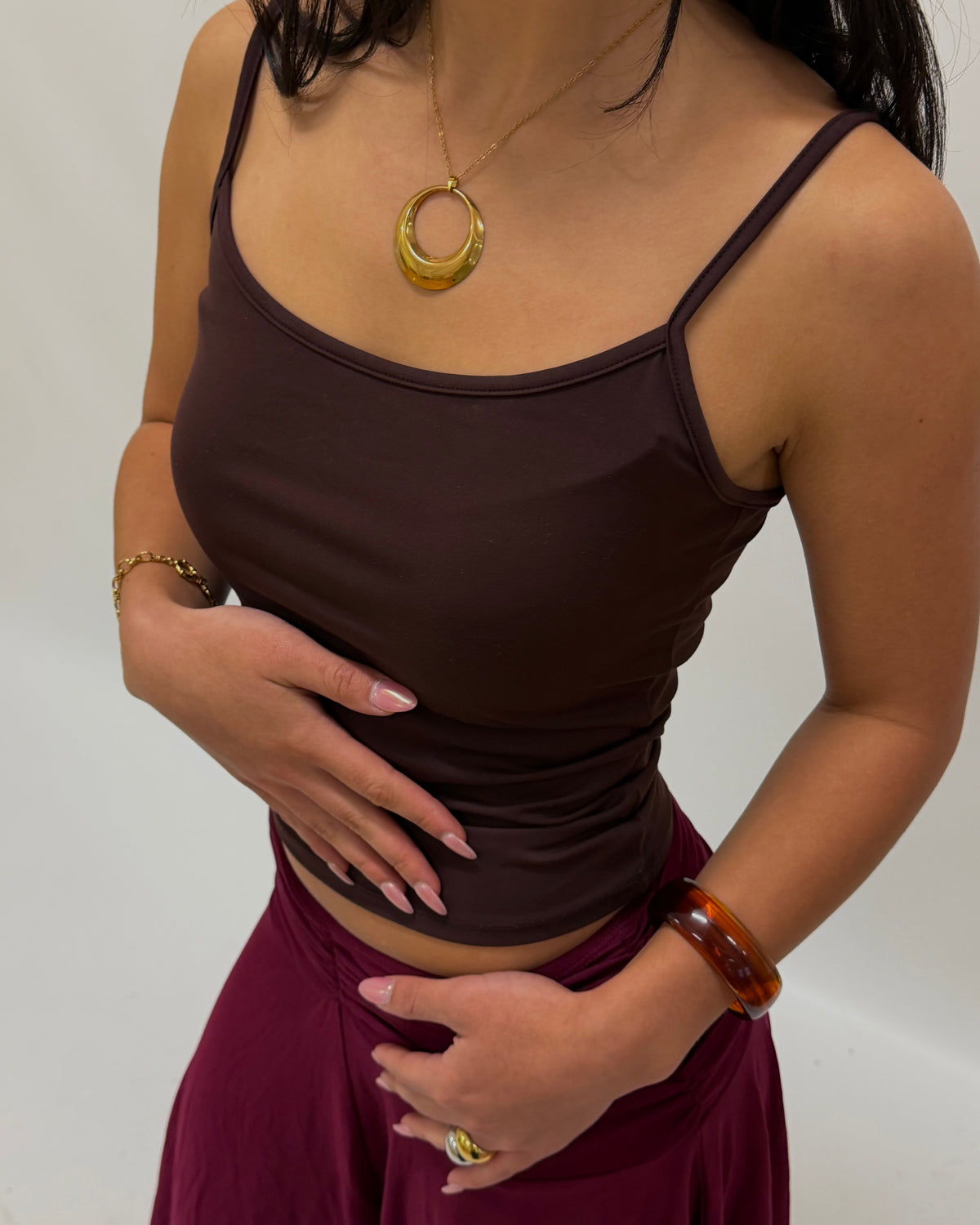 A woman wearing a dark brown spaghetti-strap camisole top and a deep magenta skirt, accessorized with chic gold jewelry including a large circular pendant, brown bangle bracelet, and rings.