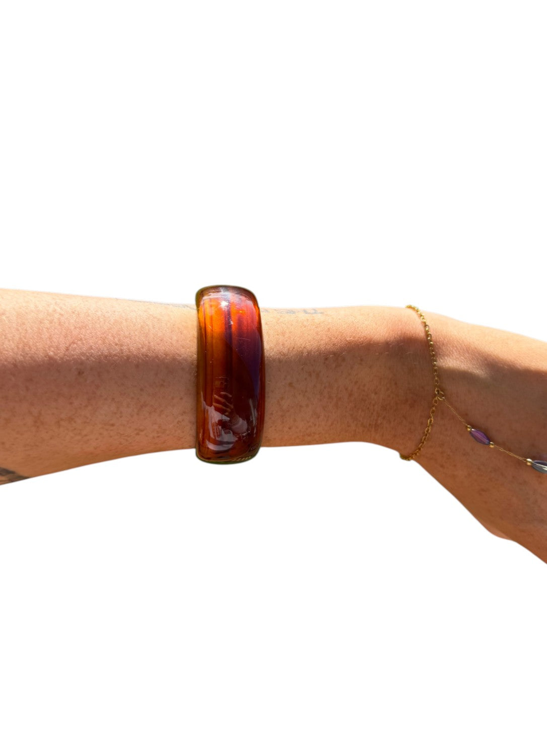 A translucent amber-brown glass bangle bracelet worn on a person's wrist, shown against a plain white background. A thin gold chain bracelet is also visible on the same wrist.