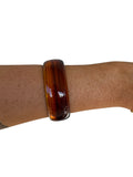 A glossy amber-brown tortoiseshell bangle bracelet worn on a person's wrist, shown against a plain white background.