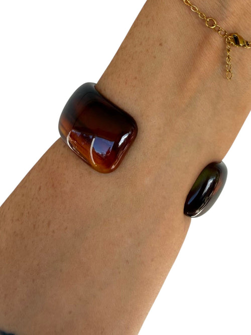 A close-up of a person's wrist wearing a chunky, glossy dark brown glass cuff bracelet, with a second matching dark oval glass bracelet and a gold chain bracelet also visible against a white background.