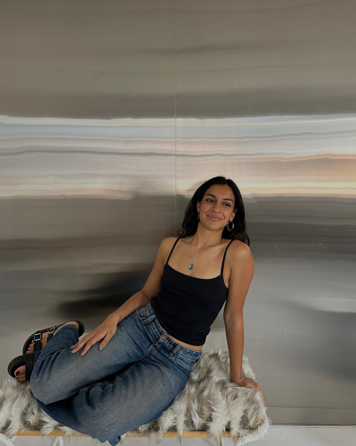 A woman wearing a black spaghetti-strap tank top and blue jeans sits on a white faux-fur rug in front of a large abstract wall art piece.