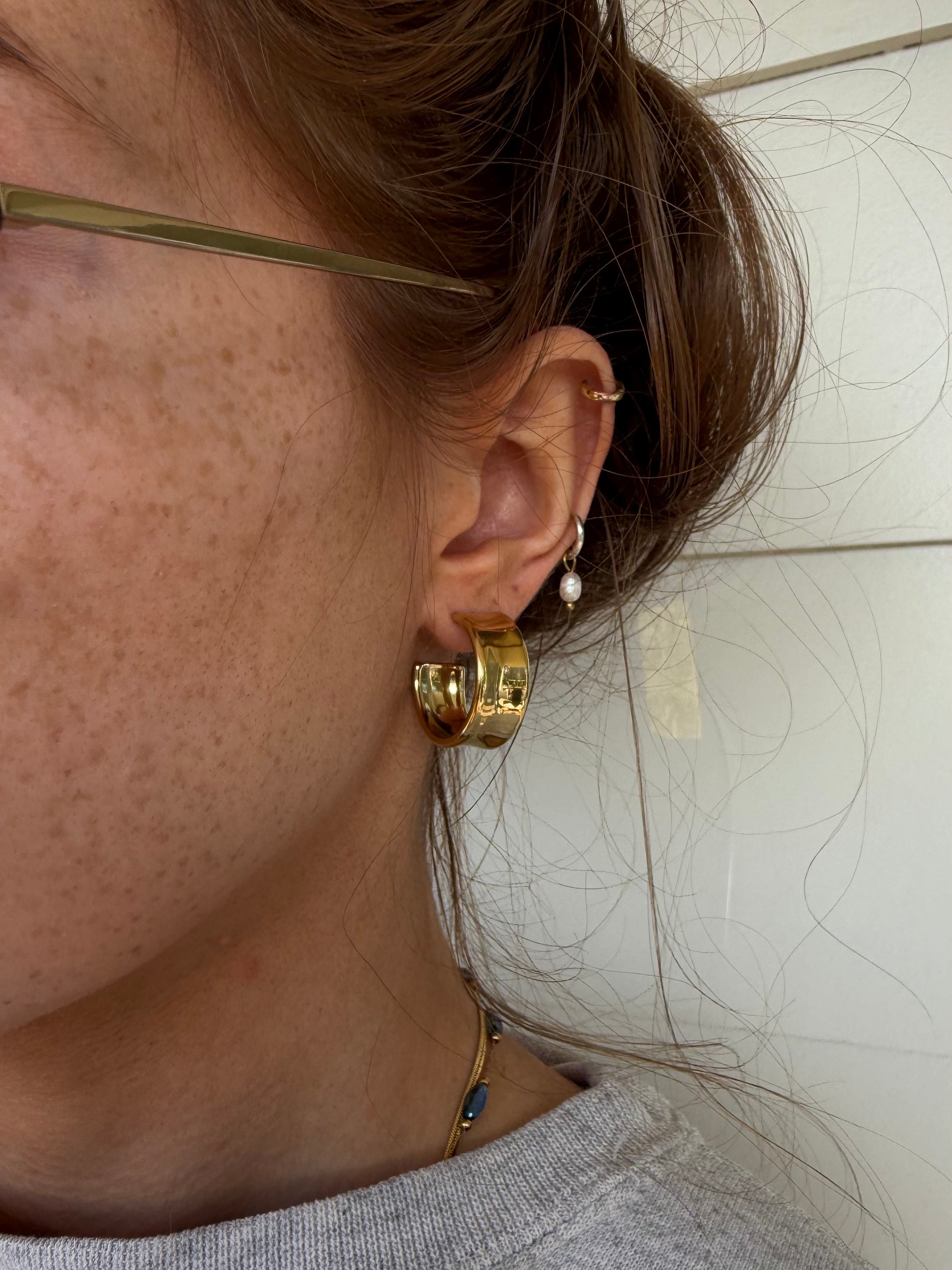 A close-up side profile of a person wearing a pair of chunky gold hoop earrings, a small gold huggie hoop, and a pearl drop earring, with brown hair pulled back and a light-colored tiled wall in the background.