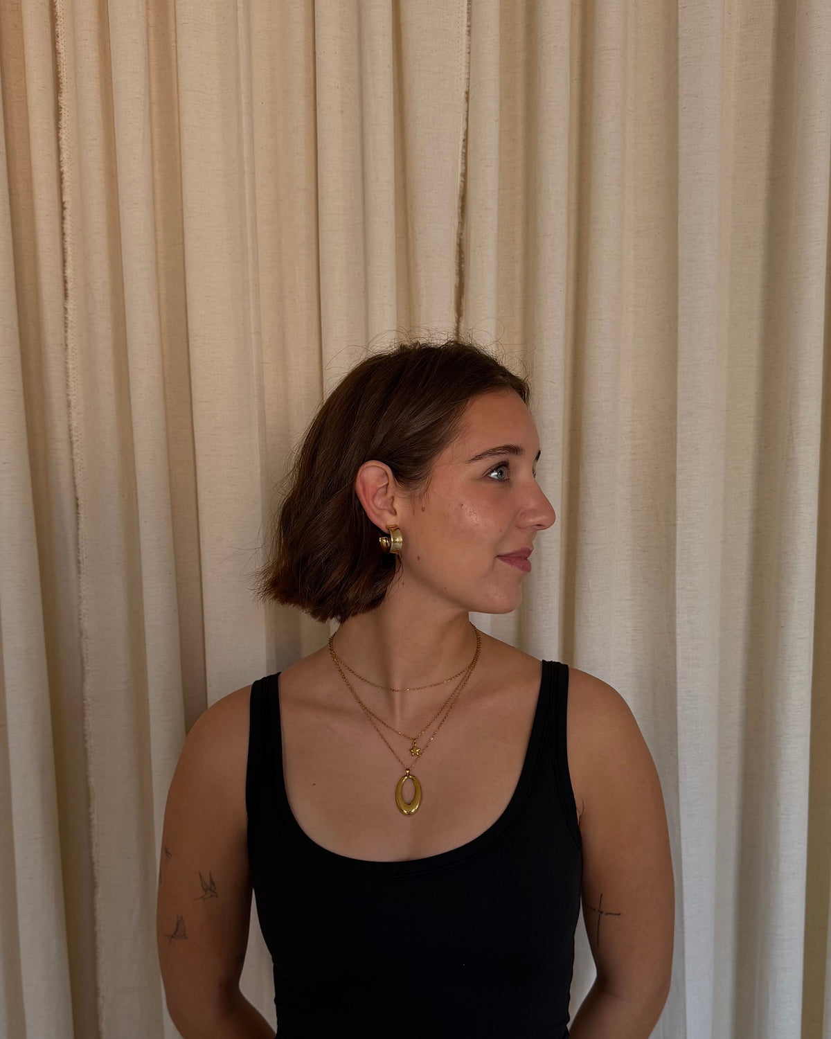 A woman with short brown hair stands in front of beige curtains, wearing a black tank top and light blue jeans.