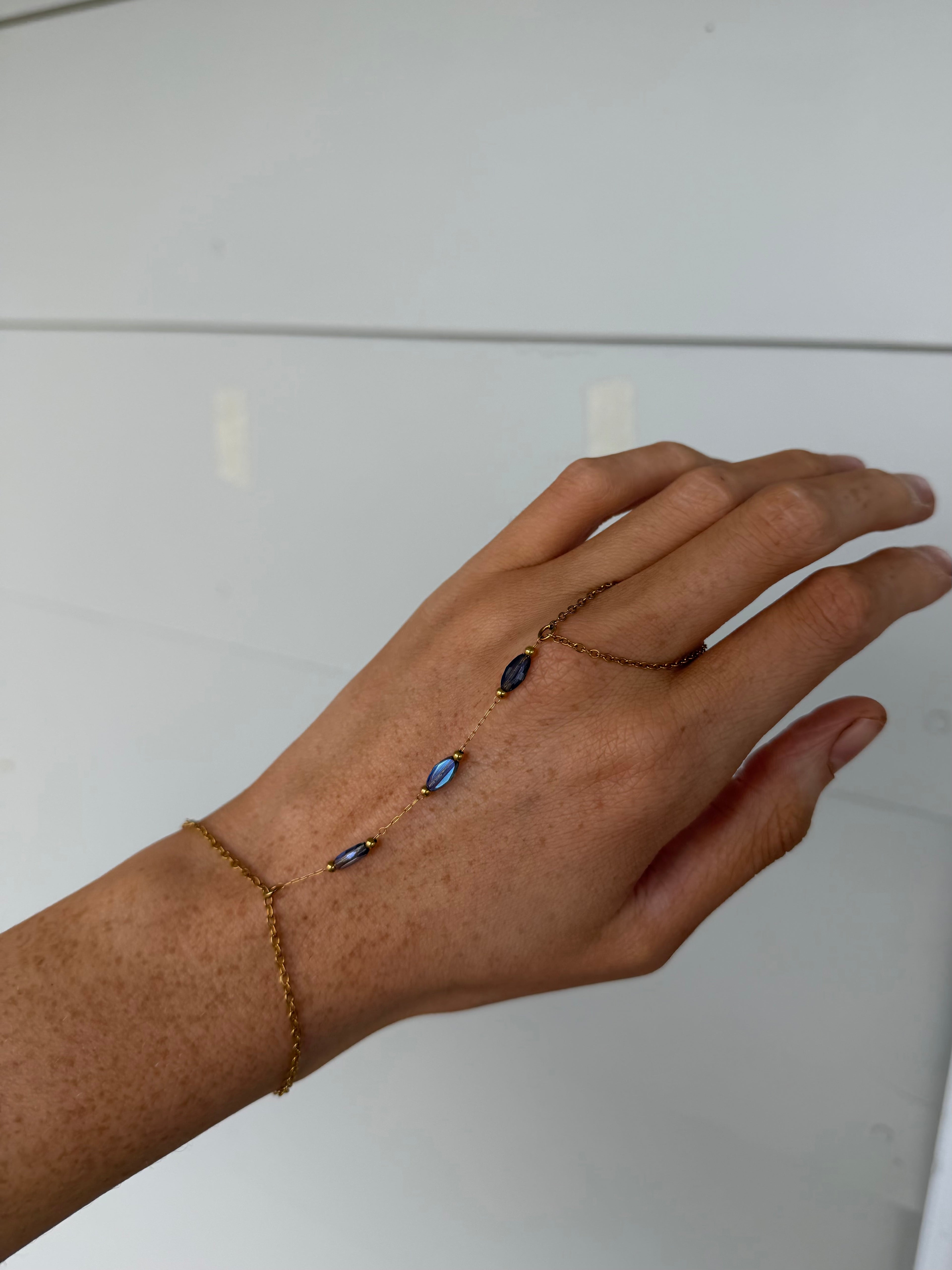 A delicate gold chain hand bracelet and matching ring worn on a person's hand, featuring small dark blue oval stones, shown against a plain light background.