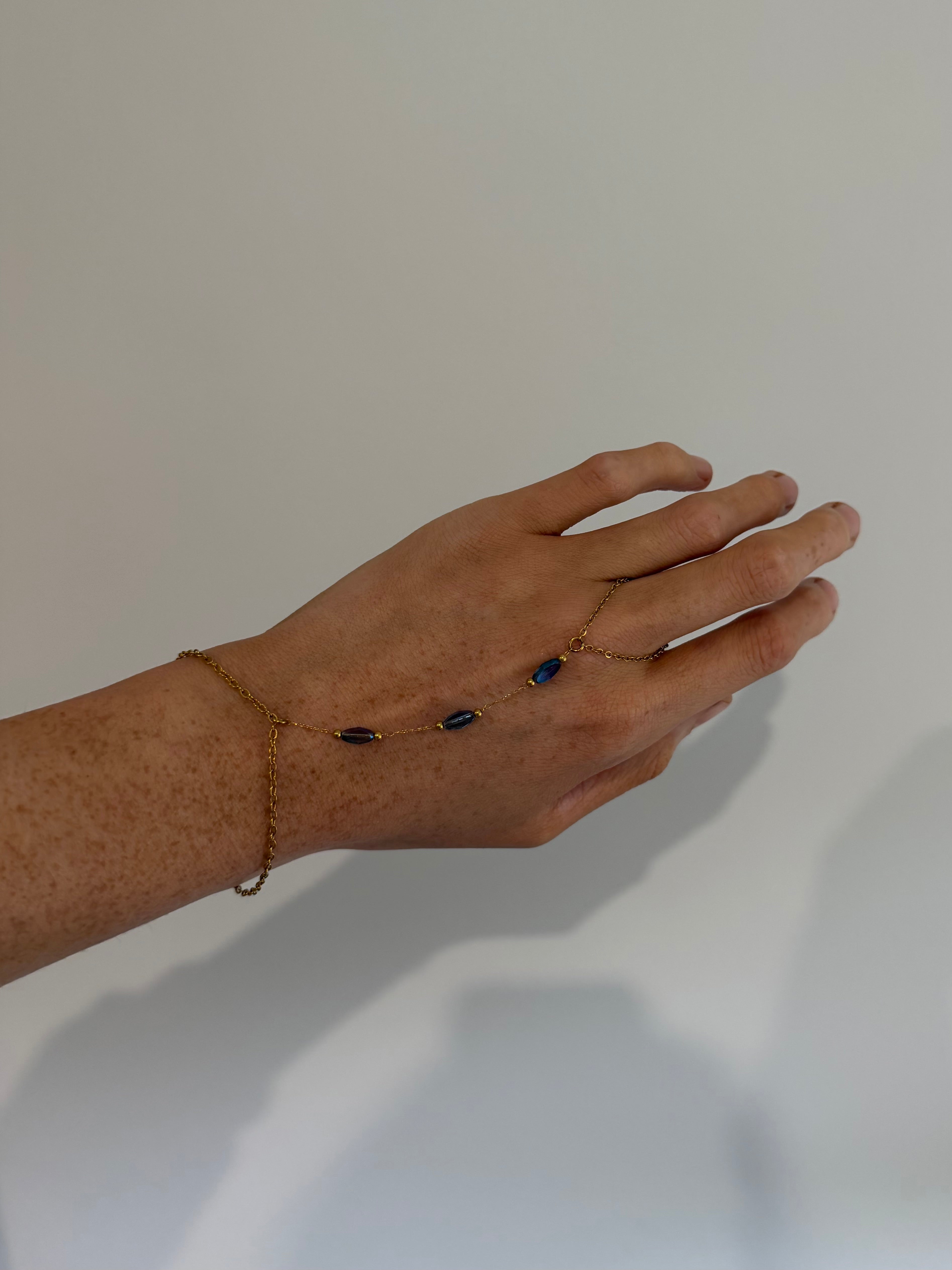 A delicate gold chain foot chain bracelet with small dark blue beads, shown worn on the back of a person's hand against a plain light gray background.