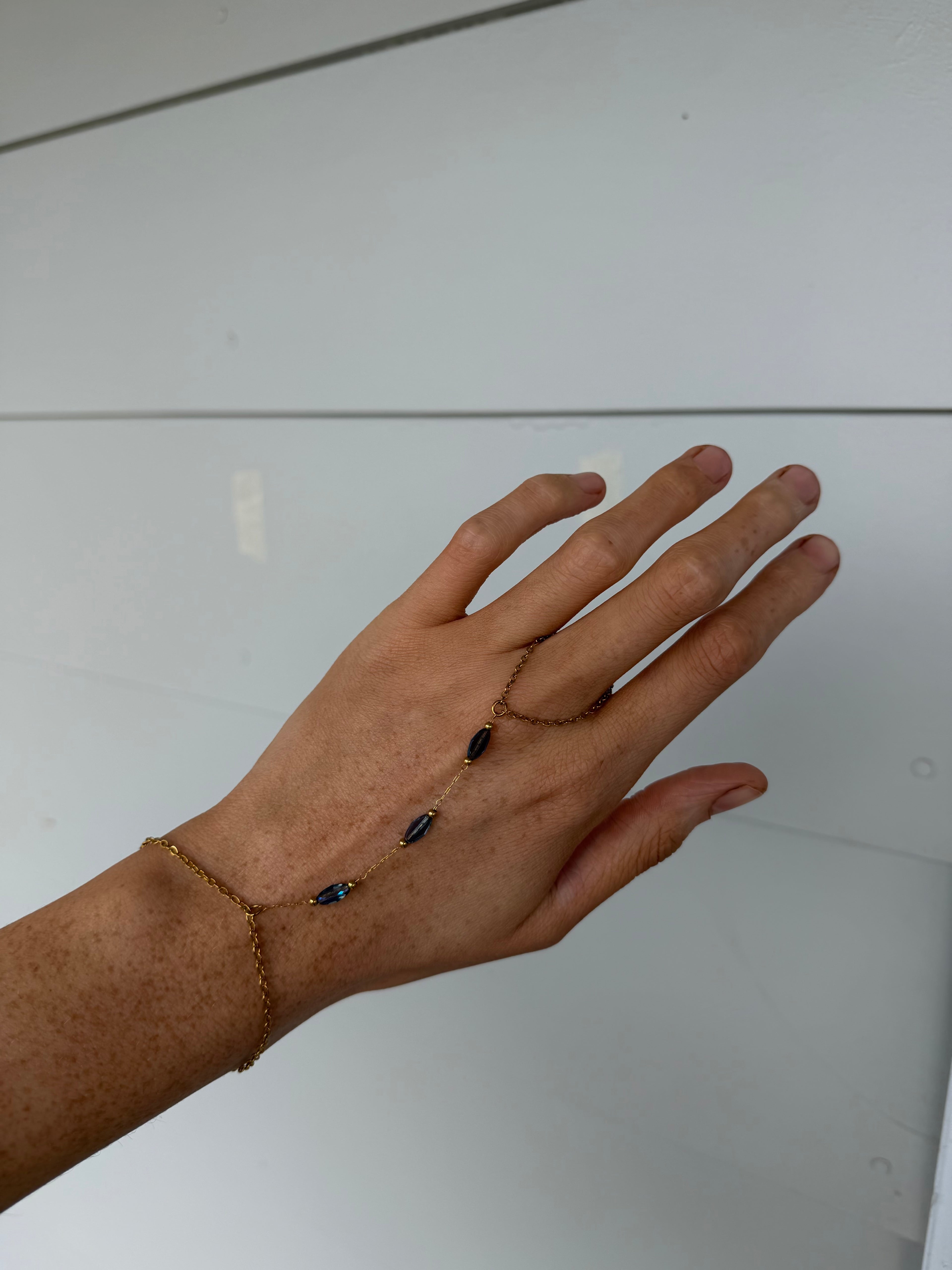 A delicate gold chain hand bracelet with three dark oval beads, worn on a person's hand against a plain light gray background.