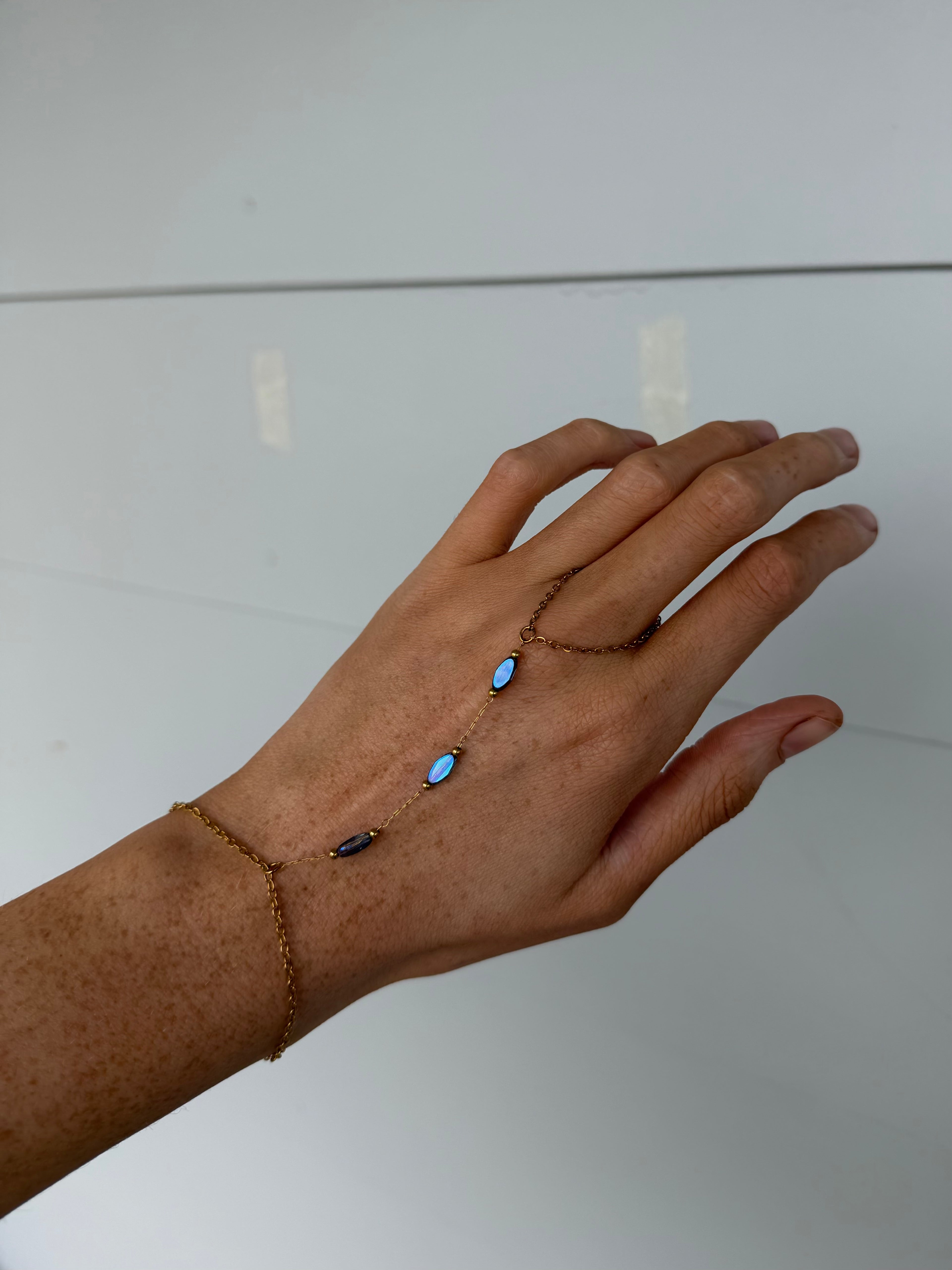 A delicate gold chain hand bracelet with three small oval blue gemstones worn on a person's hand against a plain light background.