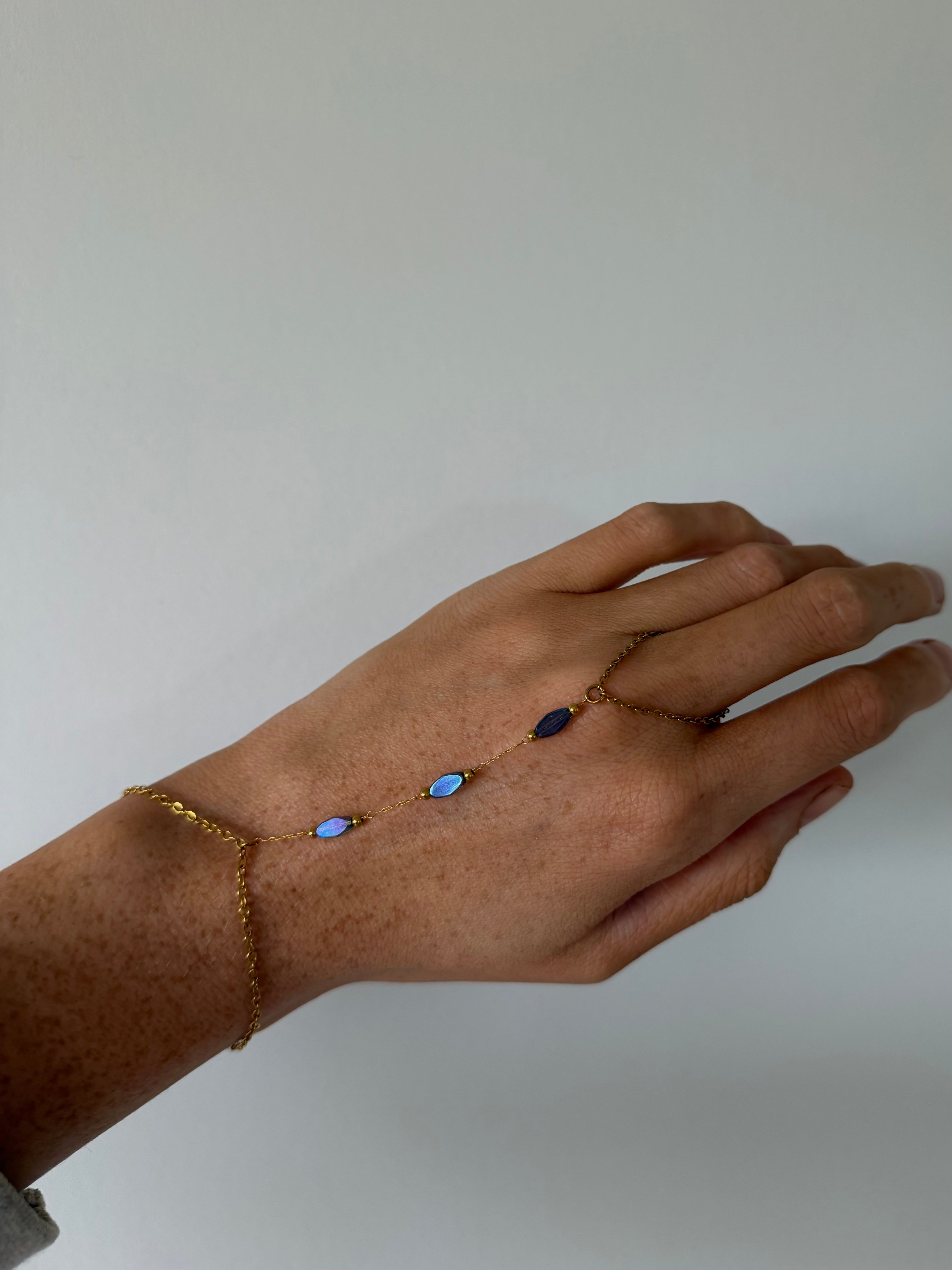 A delicate gold chain hand bracelet with three small oval blue and dark blue stones, shown worn on a person's hand against a plain light gray background.