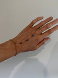 A delicate gold chain hand bracelet with three small dark blue oval stones, shown worn on a person's hand against a plain light gray background.