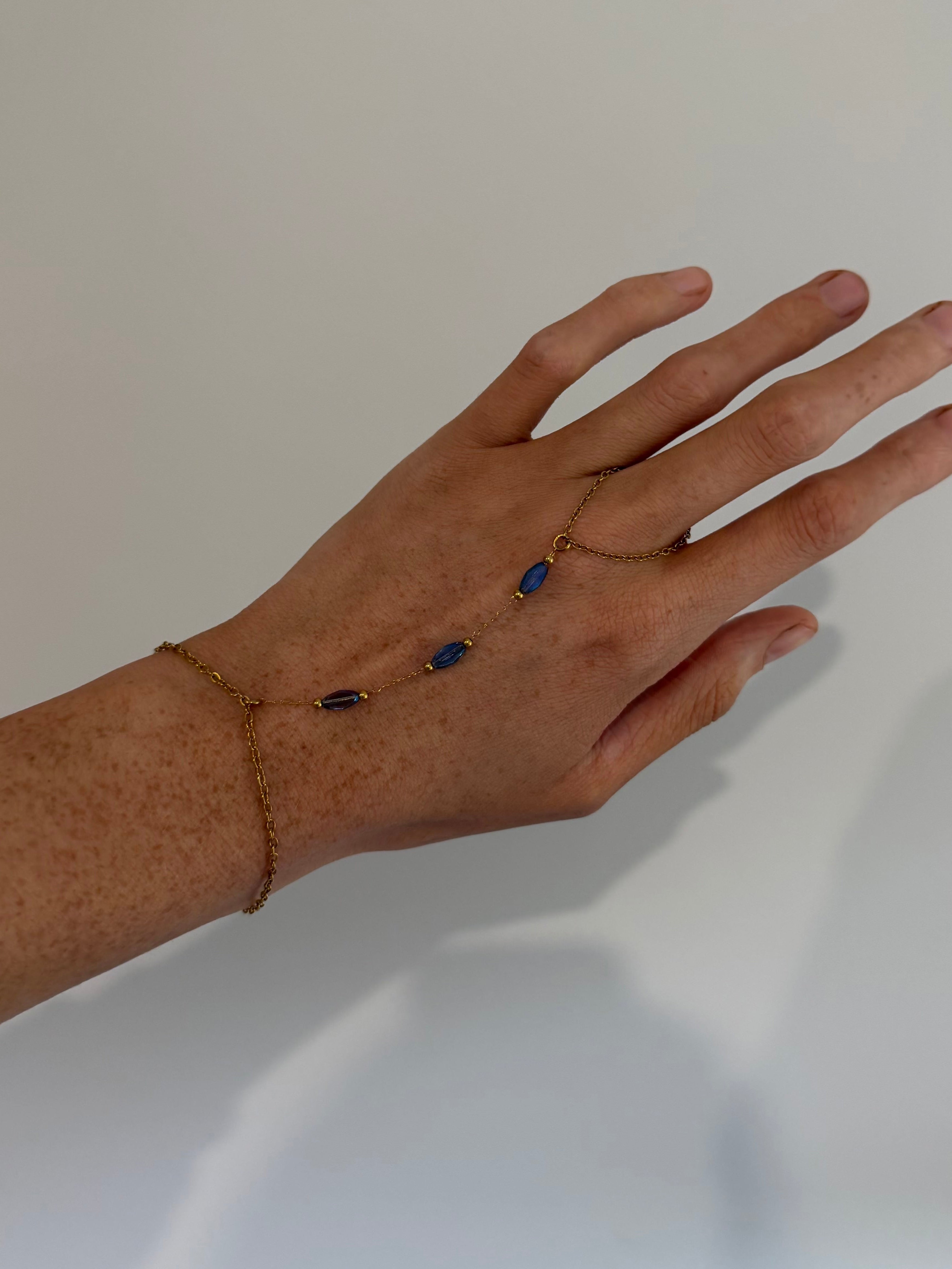 A delicate gold chain hand bracelet with three small dark blue oval stones, shown worn on a person's hand against a plain light gray background.