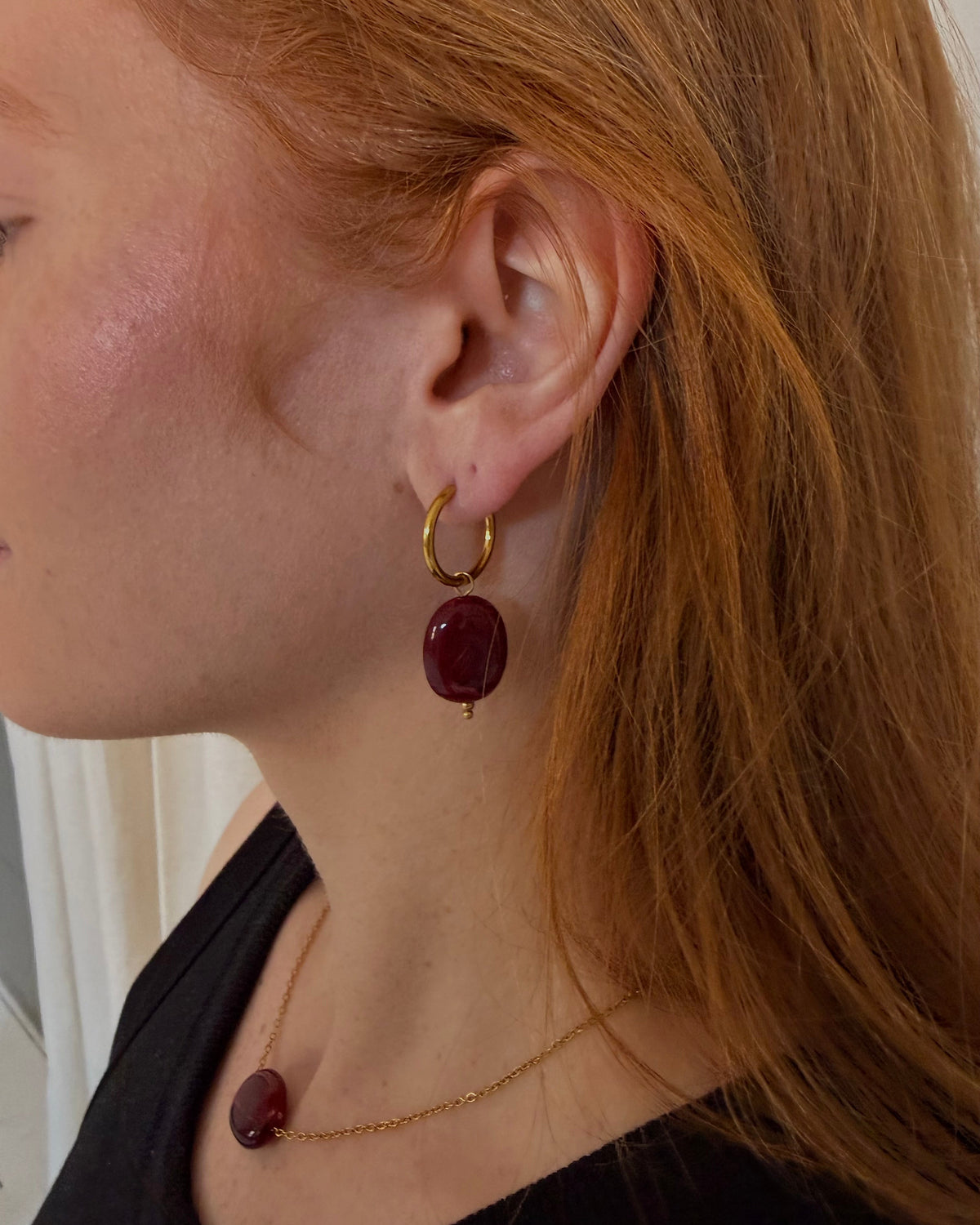 A close-up side profile of a woman with red hair wearing a gold hoop earring with a dark red oval drop and a matching gold necklace with a dark red oval drop.