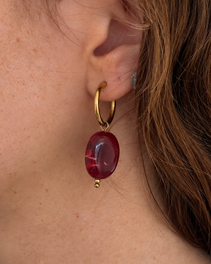 A close-up side profile of a person wearing a gold hoop earring with a polished oval dark red drop pendant.