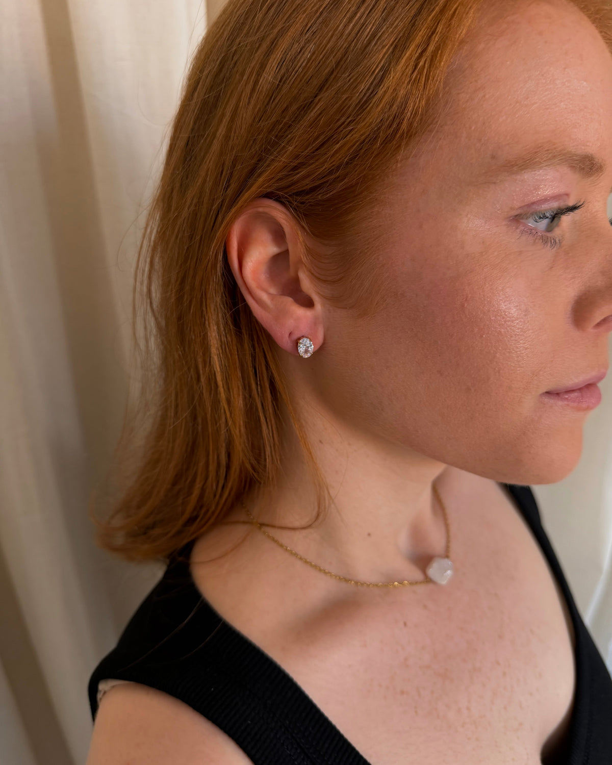A close-up side profile of a woman with red hair wearing a small round stud earring and a delicate gold necklace with a white bead, set against a soft beige curtain background.
