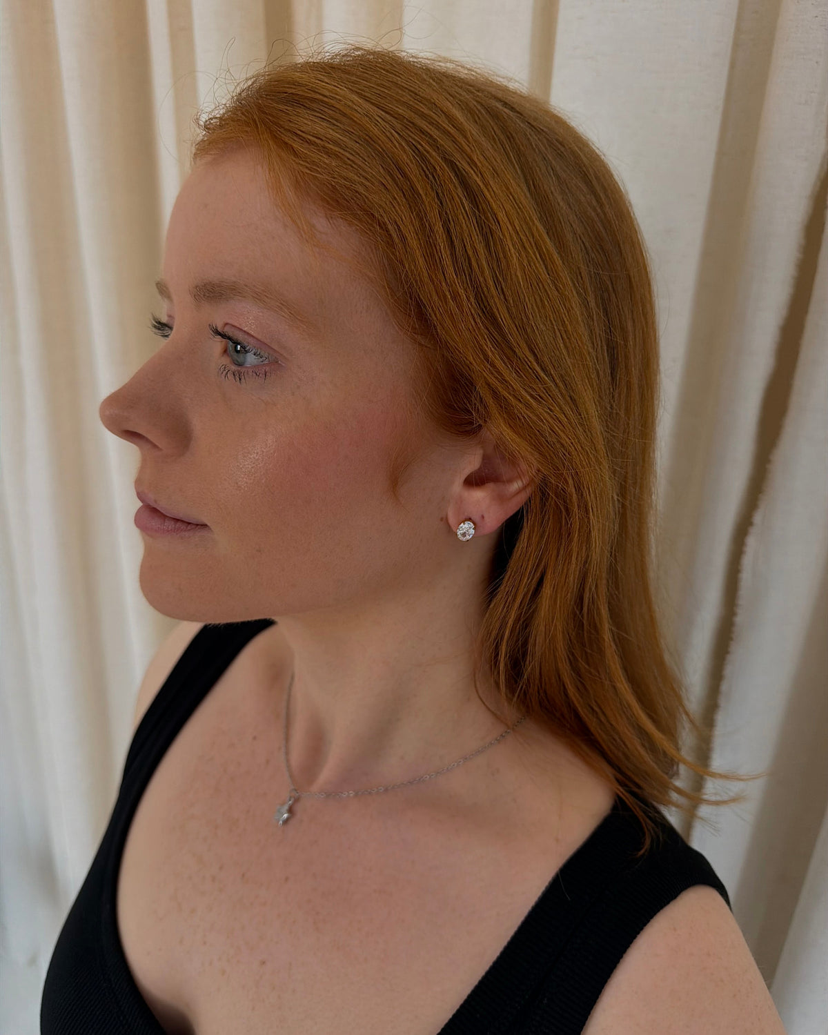 A woman with red hair is shown in profile wearing a small silver stud earring and a delicate silver necklace against a neutral cream curtain background.