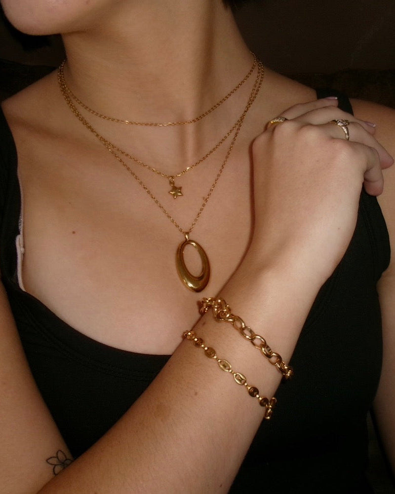 A close-up lifestyle shot of a person wearing layered gold jewelry, including a gold oval pendant necklace, a gold chain necklace with a small star charm, a gold chain bracelet, and a gold curb bracelet, while wearing a black tank top.