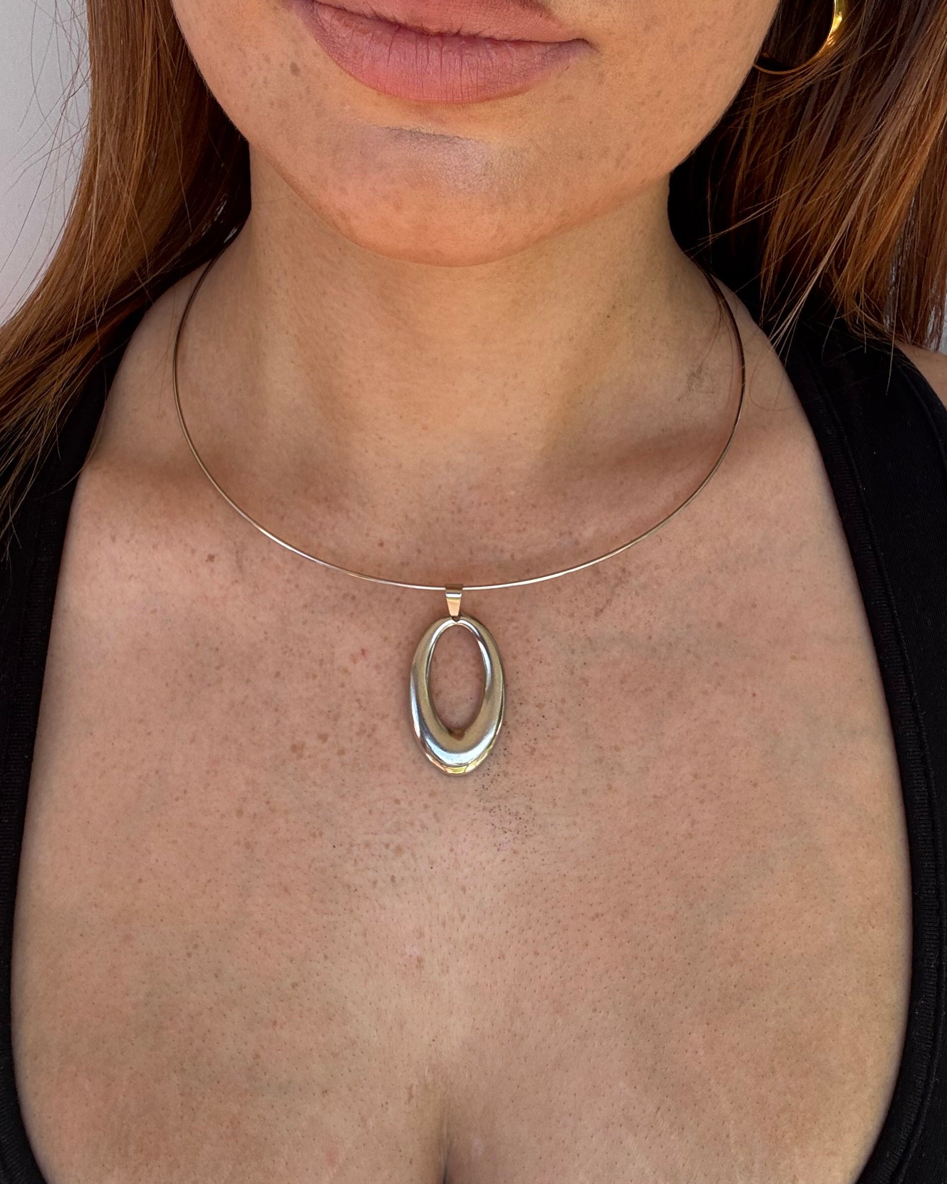 A close-up of a person lying on their back wearing a silver circular collar necklace with a central oval link and a thin circular band extending around the neck.