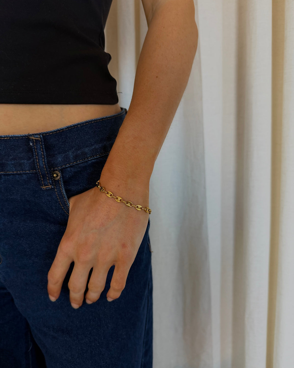 A close-up of a person wearing a delicate gold chain bracelet on their wrist, paired with a black crop top and dark blue jeans, standing in front of a light beige curtain.