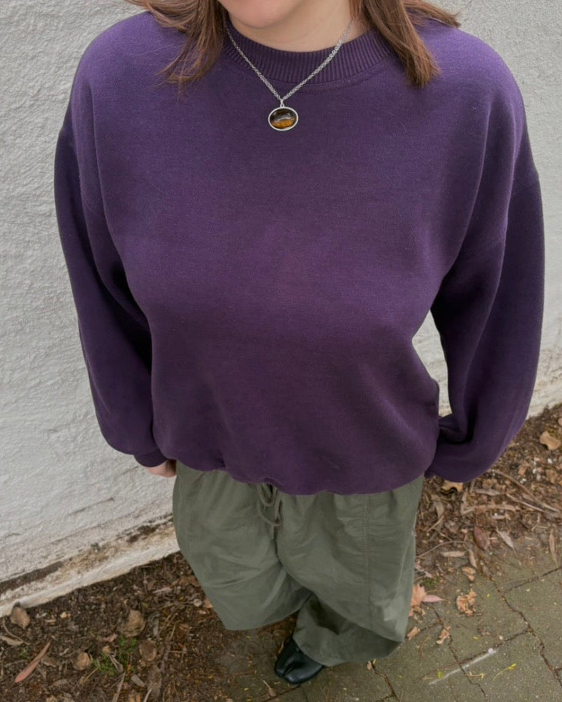 A person wearing a purple crewneck knit sweater and olive green pants, standing outdoors against a textured white wall.