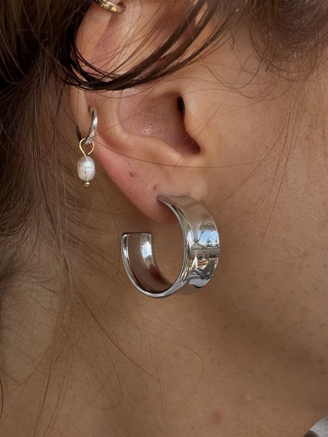 A close-up of a person's ear showcasing a chunky, polished silver huggie hoop earring, with a smaller gold hoop and white pearl drop earring also visible.