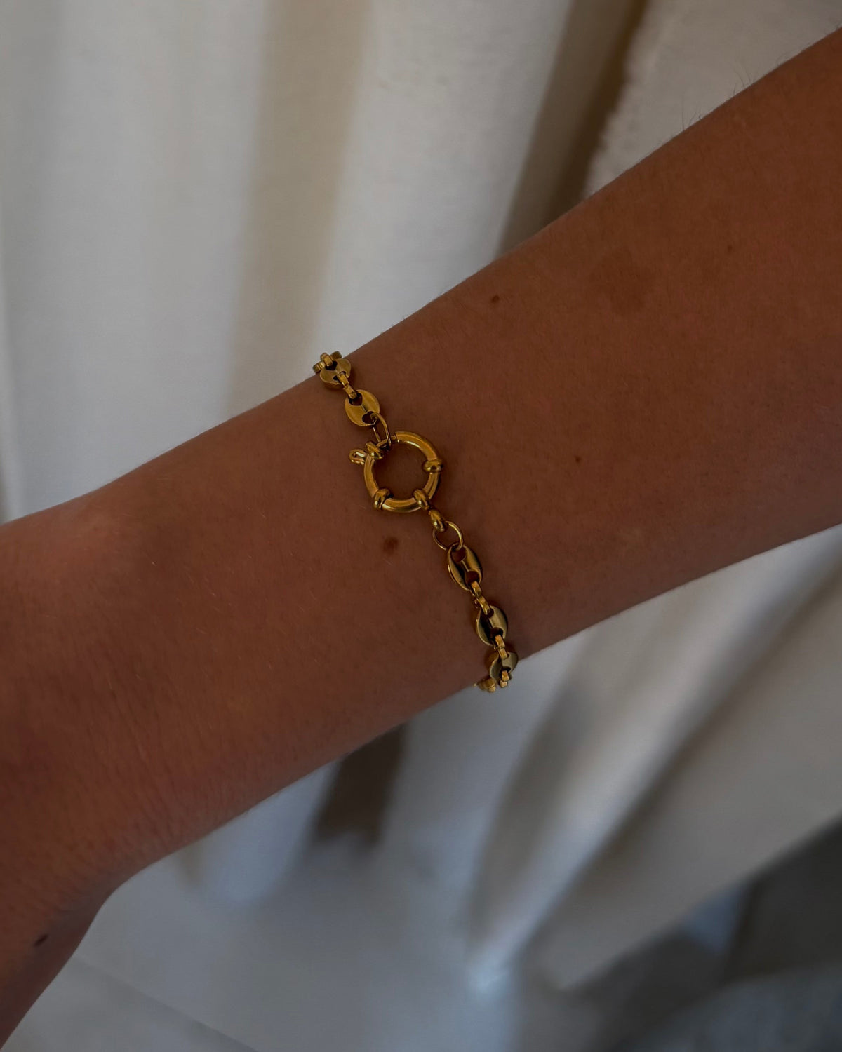 A gold chain bracelet with a small interlocking circular charm worn on a person's wrist, shown against a background of draped off-white curtains.