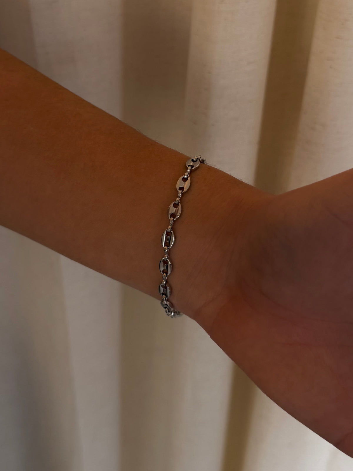 A close-up of a person's wrist wearing a delicate silver chain link bracelet, set against a soft beige curtain background.