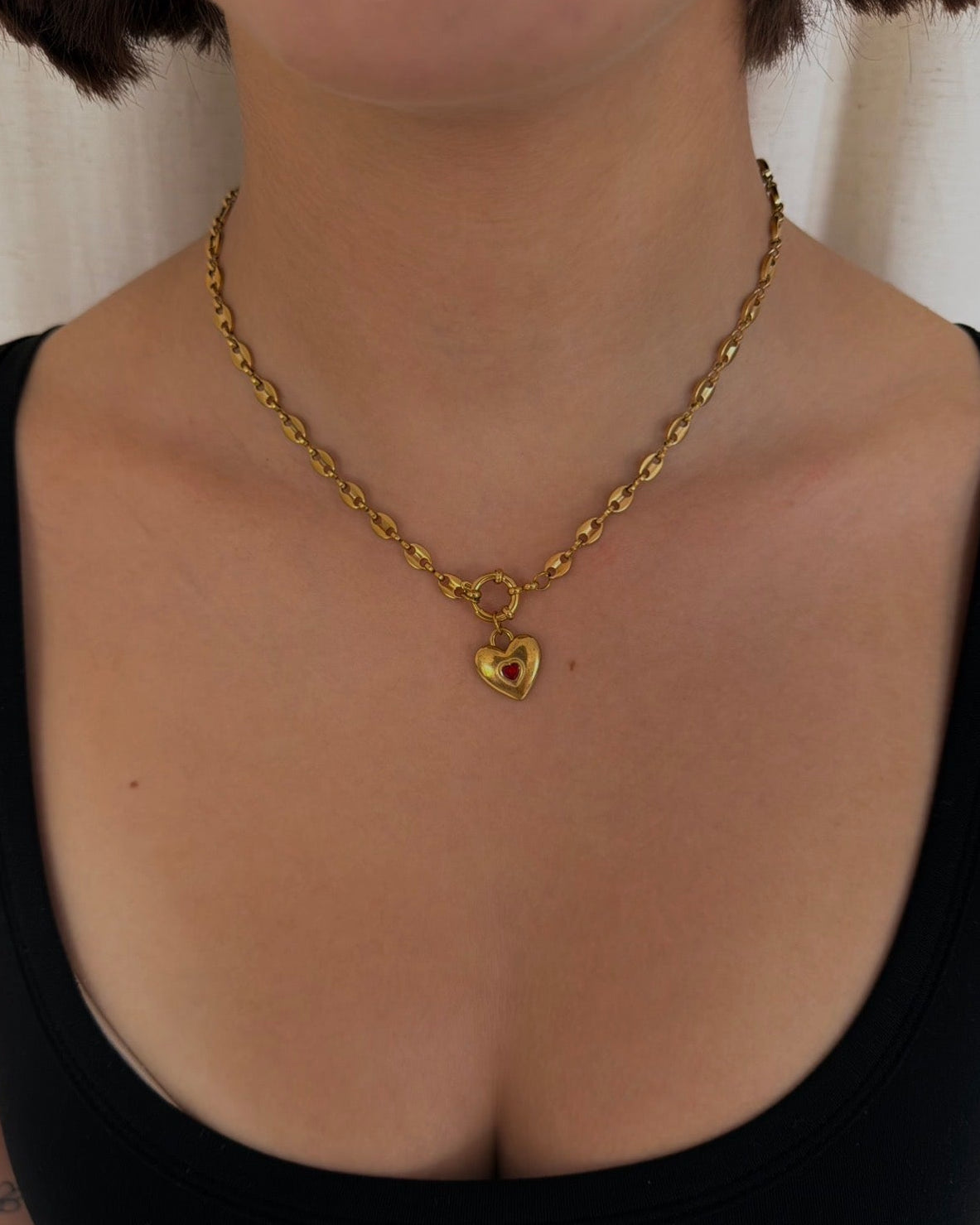 A close-up of a person wearing a delicate gold chain necklace with a small heart-shaped charm, styled with a black tank top.
