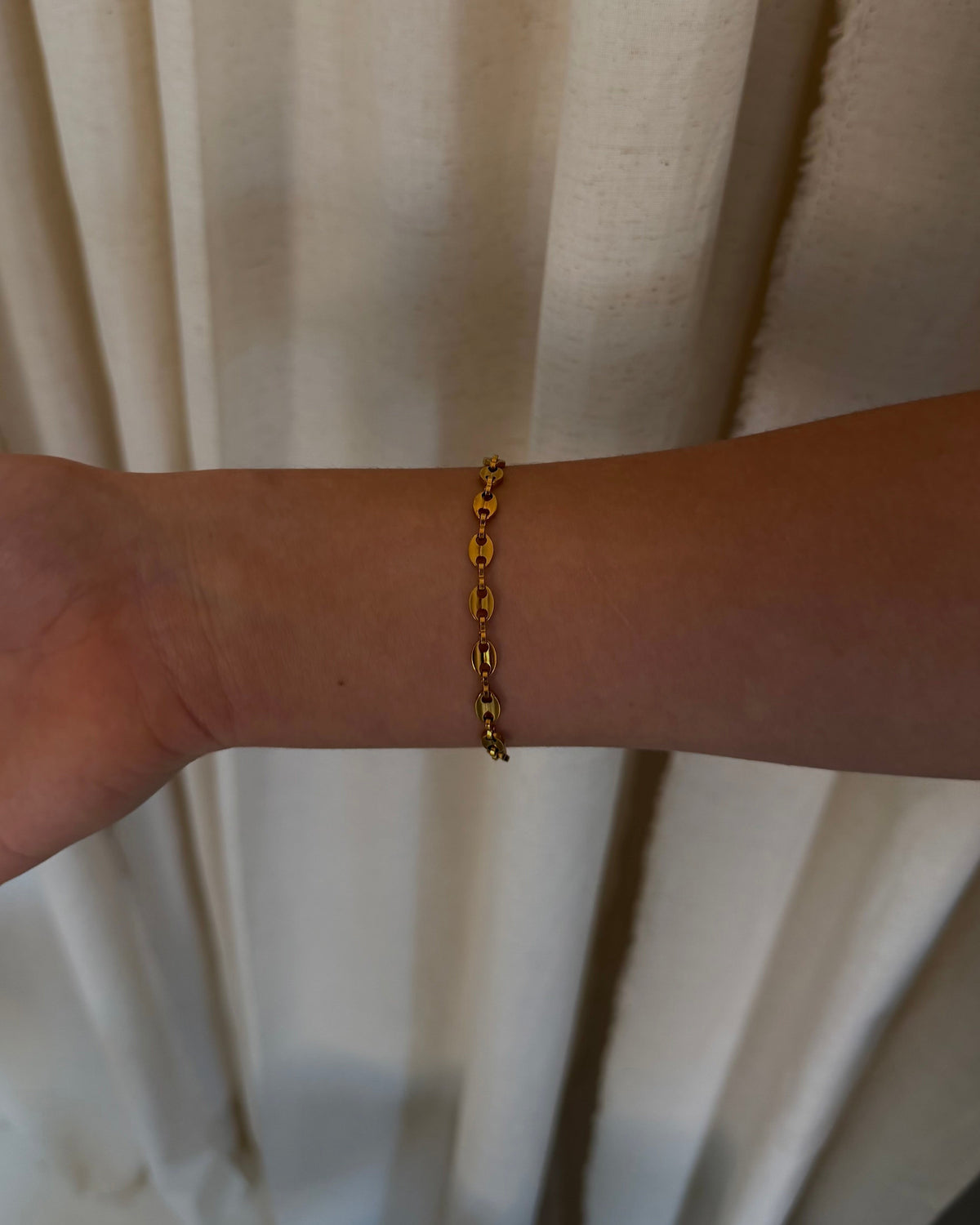A gold chain bracelet with small heart-shaped links worn on a person's wrist, shown against a background of beige curtains.