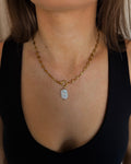 A close-up of a woman wearing a gold chain necklace with a small white shell pendant, paired with a black tank top and gold hoop earrings.