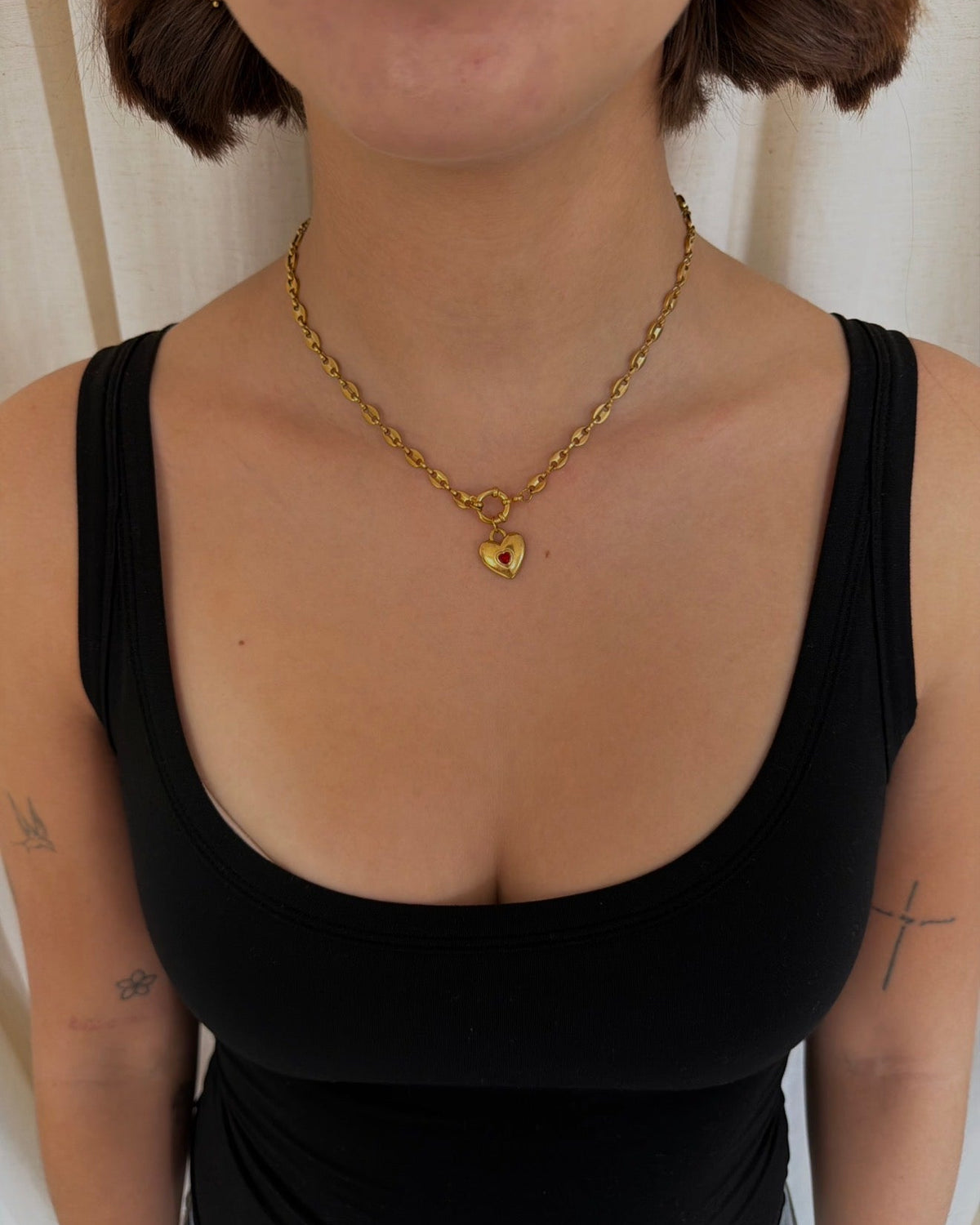 A close-up of a woman wearing a delicate gold chain necklace with a small heart-shaped pendant, paired with a black tank top.