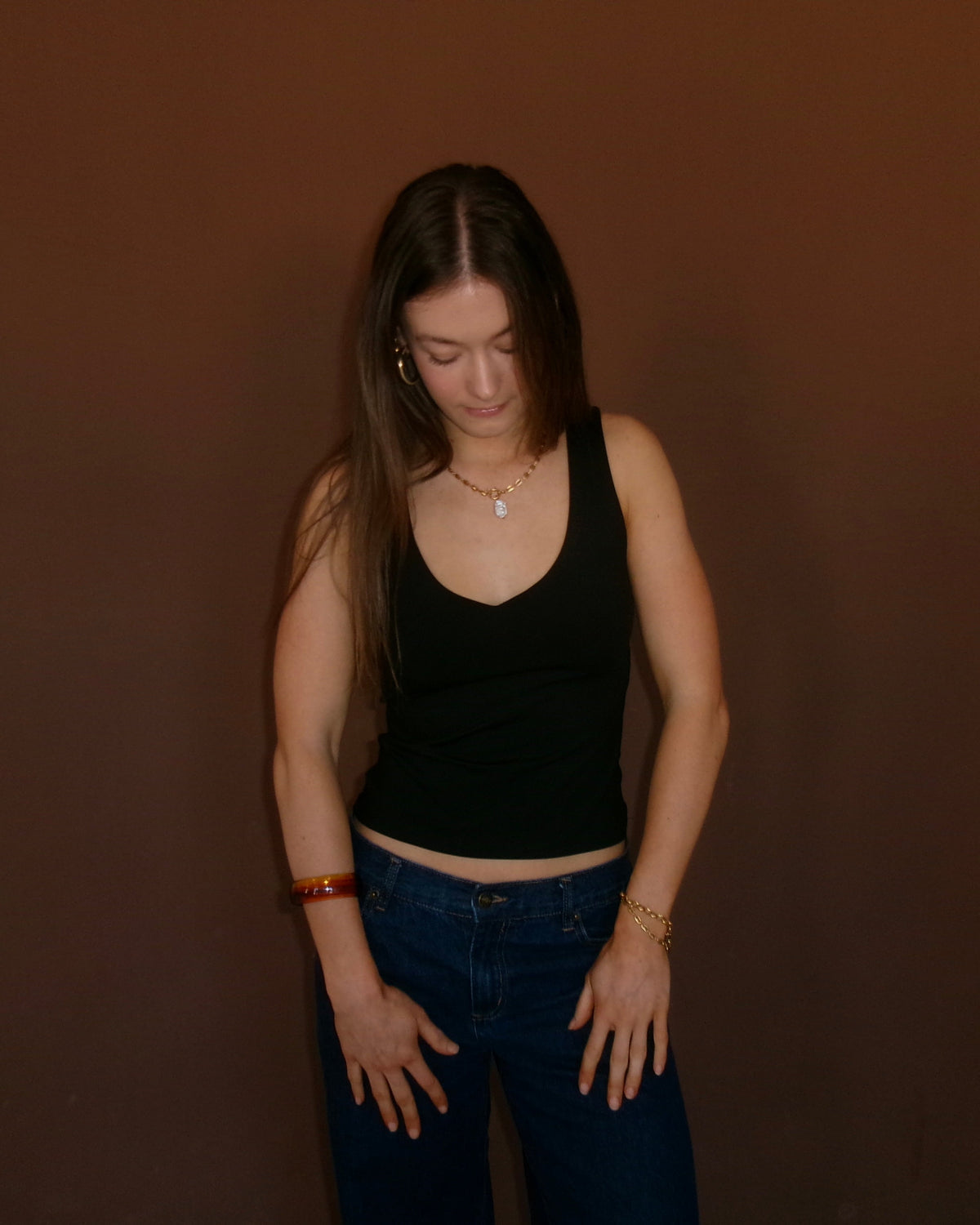 A woman with long dark hair stands against a solid dark brown background, wearing a black ribbed tank top and blue jeans.