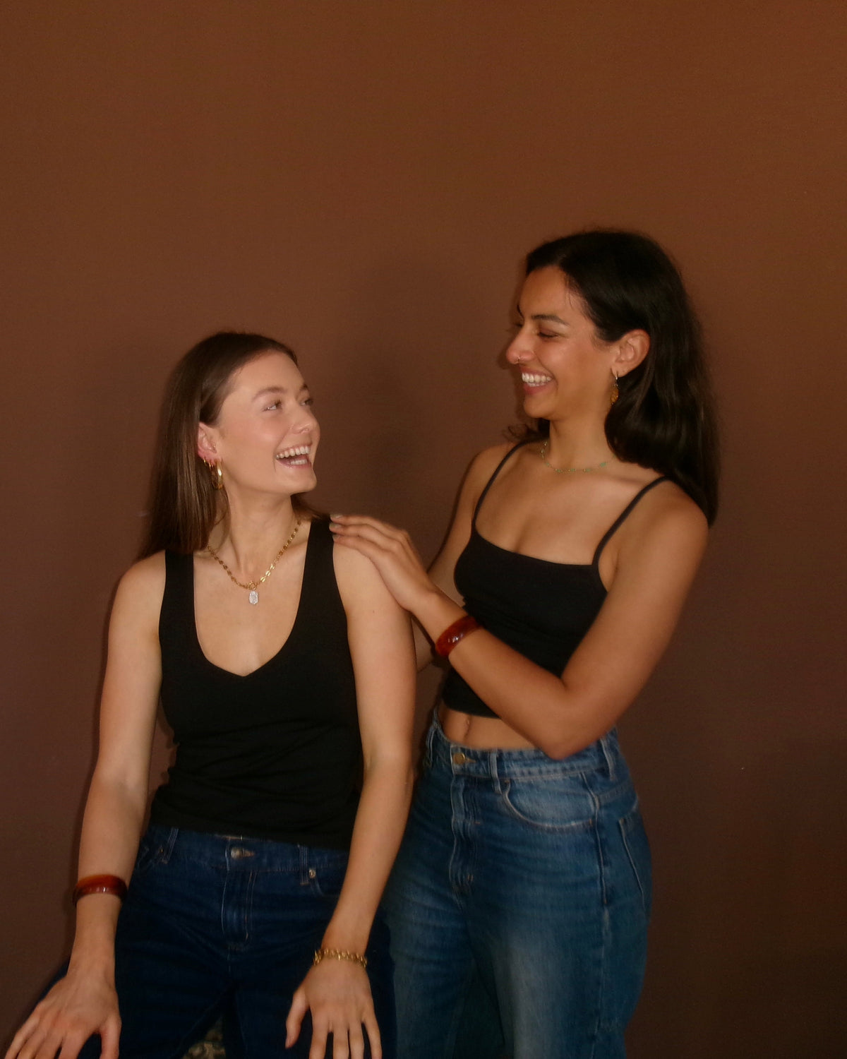 Two young women smiling and laughing while posing together against a solid dark brown wall. The woman on the left wears a black tank top and blue jeans, and the woman on the right wears a black spaghetti-strap crop top and high-waisted blue jeans.