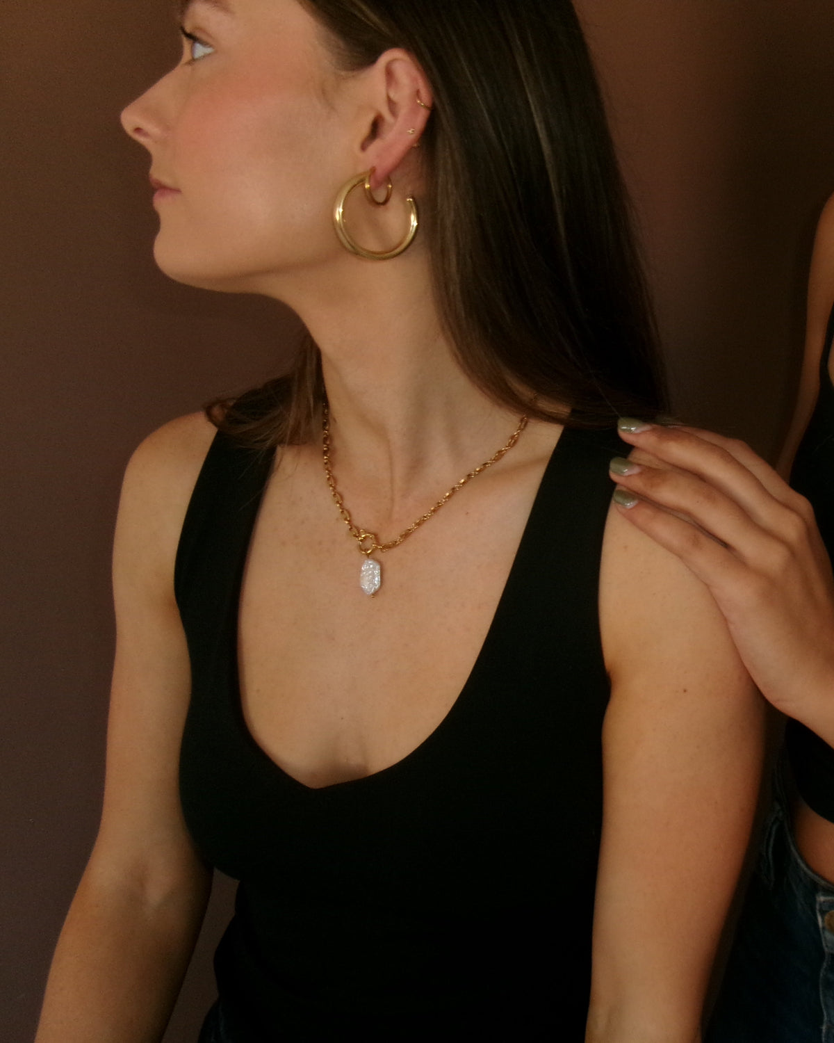 Two women wearing black camisole tops and jewelry, with one placing a hand on the other's shoulder. Visible jewelry includes a gold hoop earring, a gold pendant necklace, and a turquoise beaded necklace.
