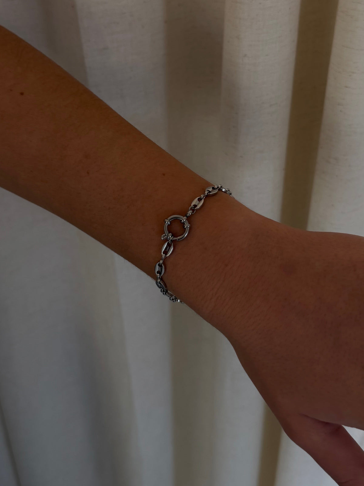 A silver-toned chain bracelet with a central circular link, worn on a person's wrist against a draped cream fabric background.