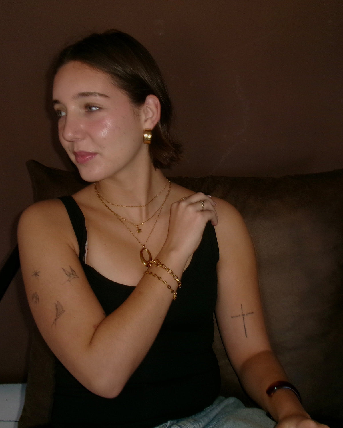 A woman with tattoos sits against a dark brown pillow and wall, wearing a black tank top and layered gold necklaces and bracelets.