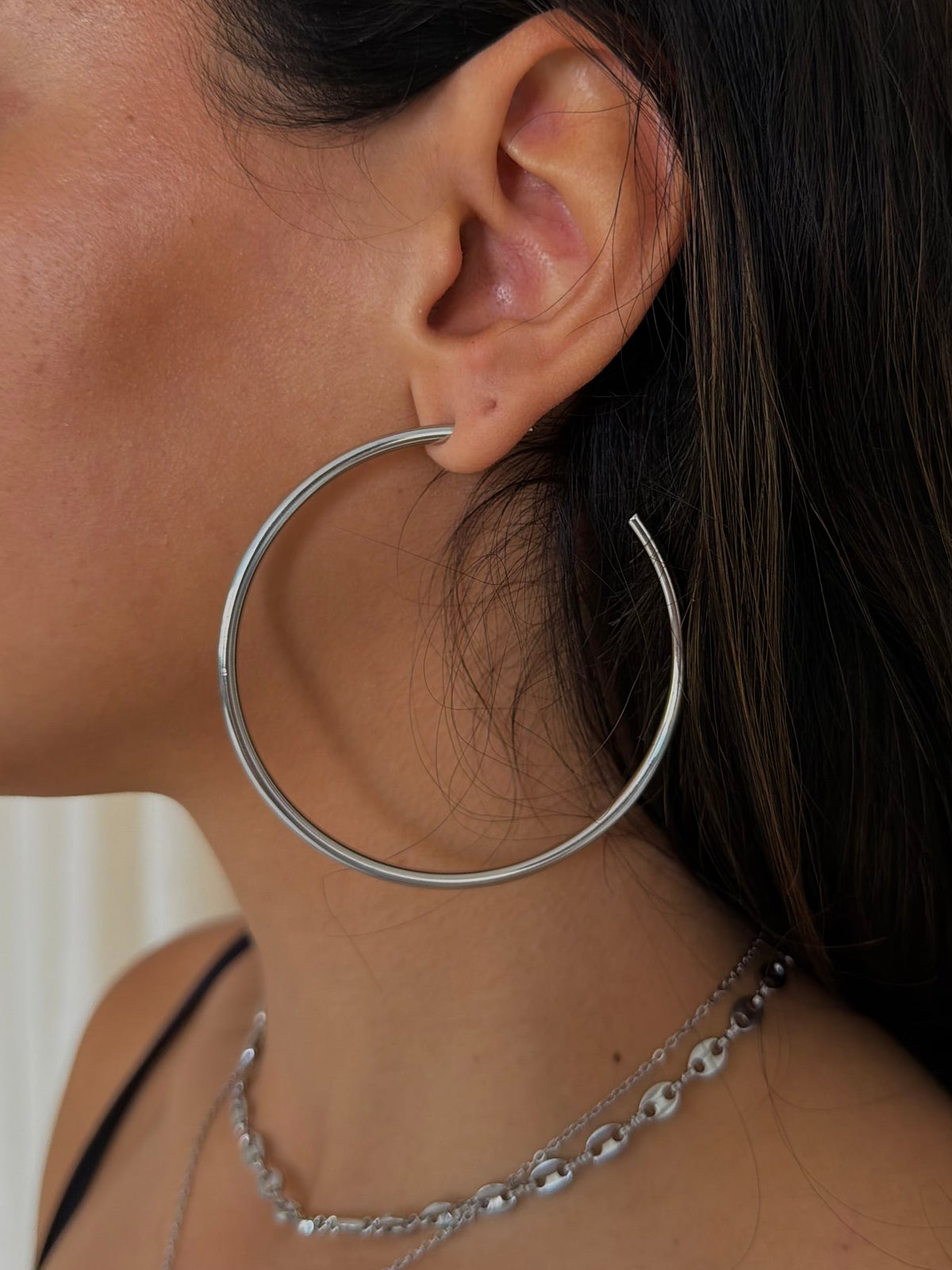 A close-up side profile of a woman wearing a large, thin silver hoop earring and layered silver necklaces.