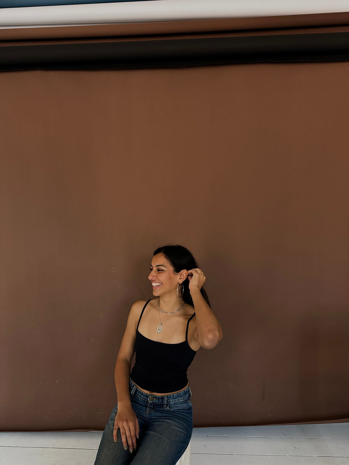 A woman wearing a black spaghetti-strap tank top and blue jeans poses against a solid brown backdrop.