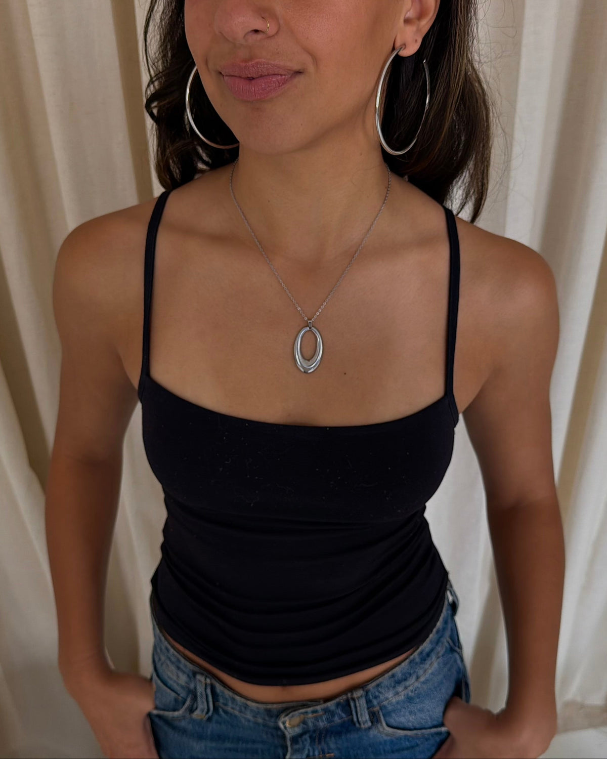 A woman wearing a black spaghetti-strap camisole top and blue jeans, standing in front of cream-colored draped curtains. She is also wearing silver hoop earrings and a silver pendant necklace.