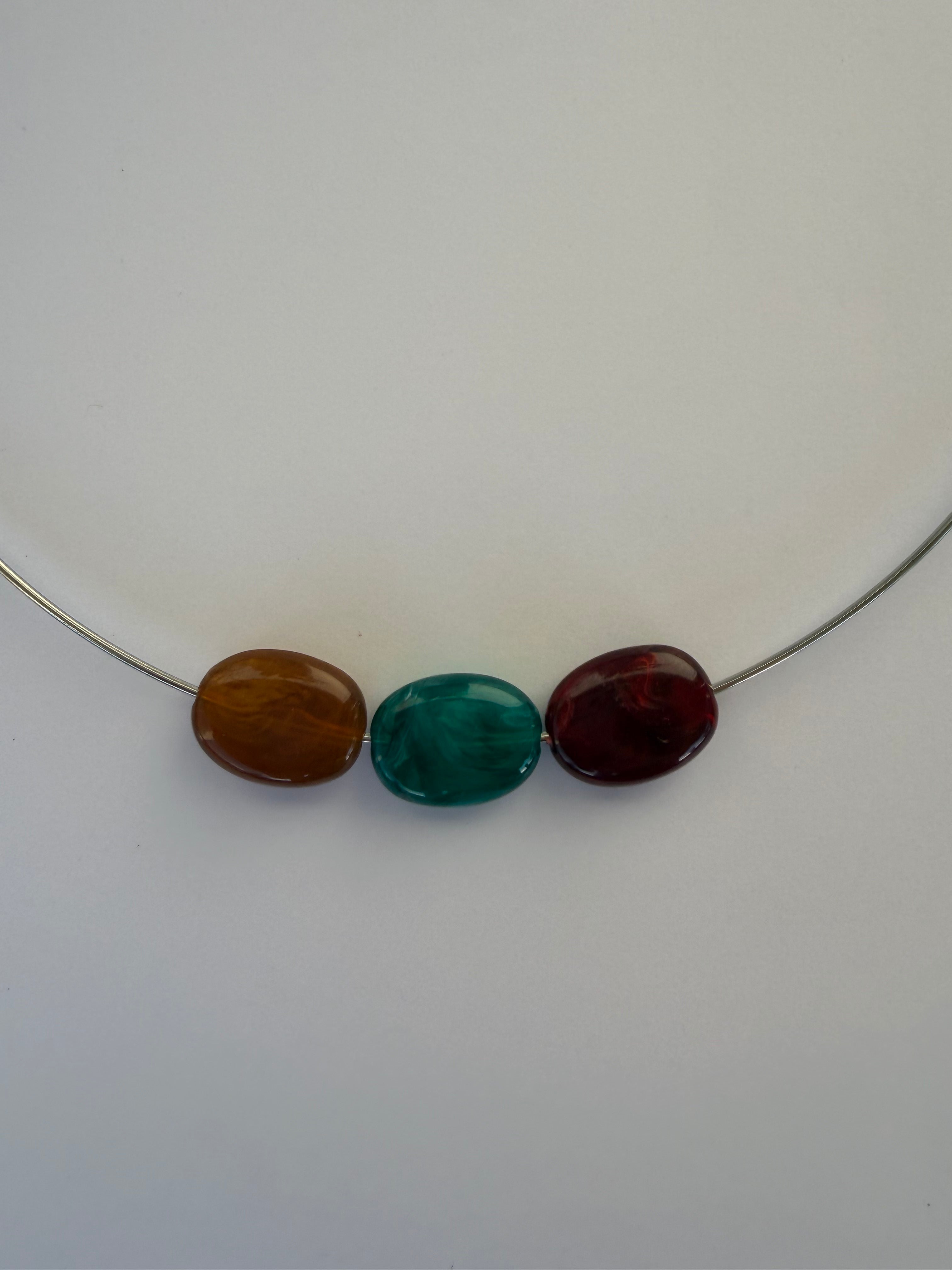 Three polished oval beads in dark red, green, and brown arranged vertically on a thin silver wire against a plain light background.