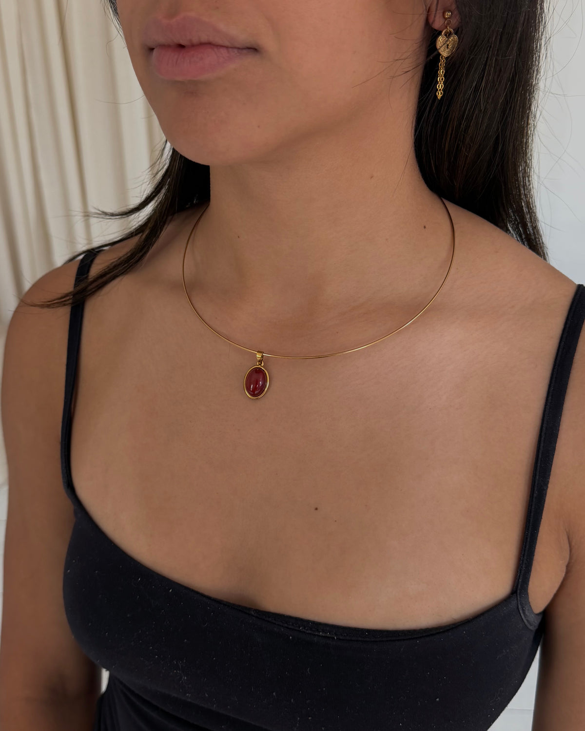 A close-up of a woman wearing a delicate gold necklace with an oval red gemstone pendant, paired with a black spaghetti-strap top.