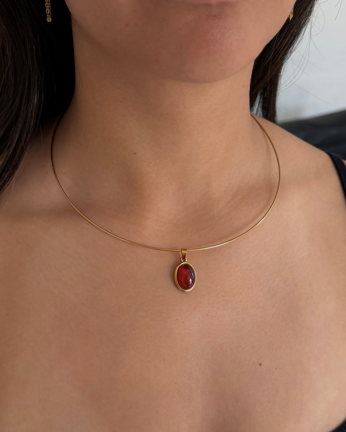 A close-up of a model's neck and chest wearing a delicate gold chain necklace with an oval red gemstone pendant.